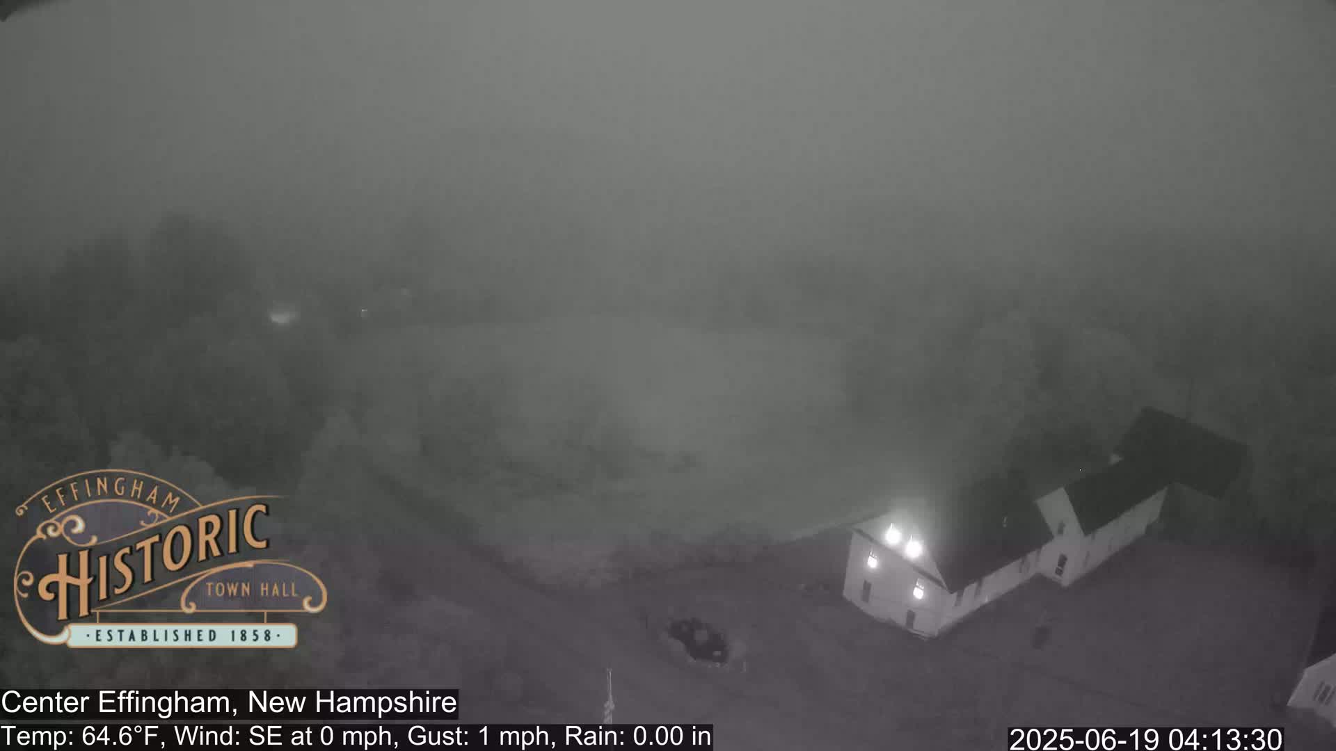 A foggy, nighttime view shows a building with lights on, and surrounding vegetation, under calm conditions.