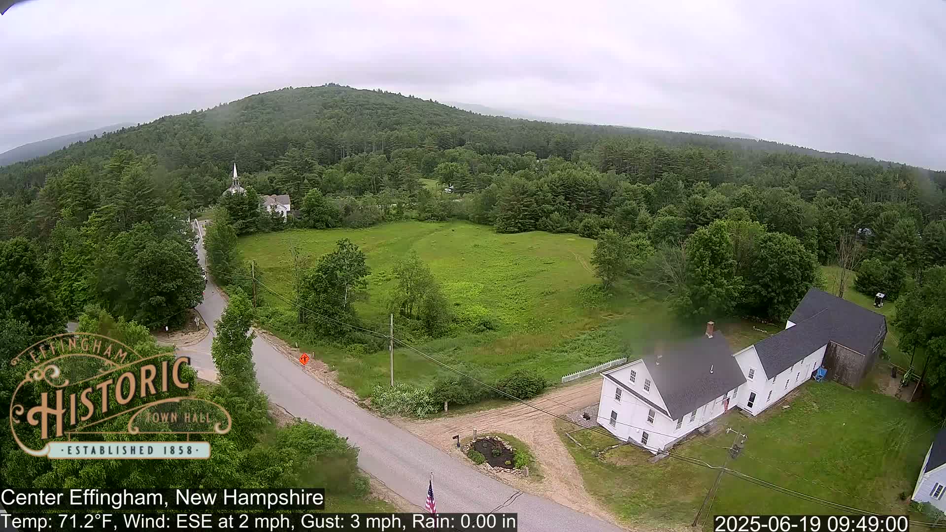 An aerial view shows a rural town nestled among green hills and trees under an overcast sky.