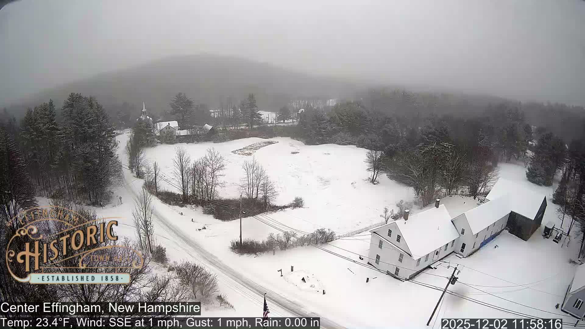 An aerial shot depicts a snow-covered village nestled among hills and bare trees, featuring a church steeple, a winding road, and several houses under a gray, overcast sky during a cold winter day.