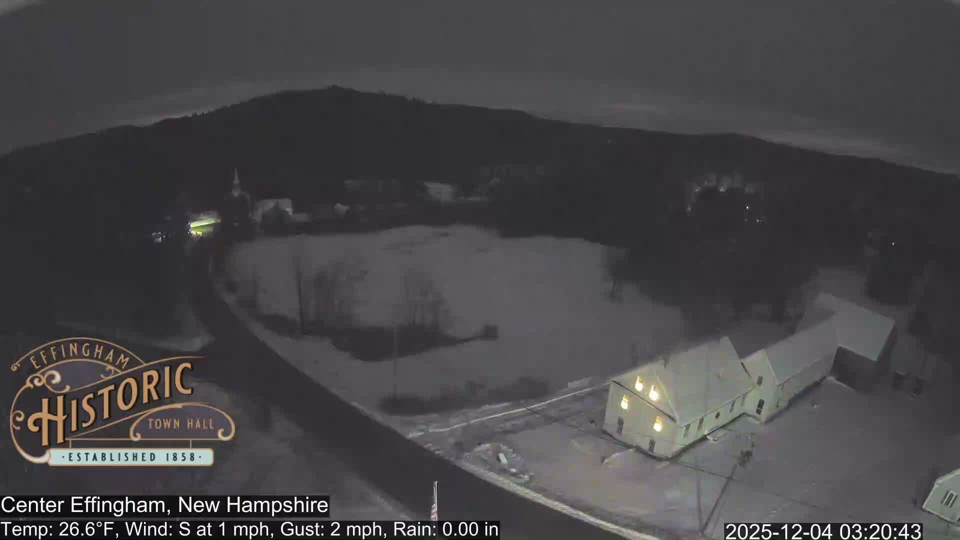 A high-angle view depicts a snow-covered village under an overcast sky during a cold winter night or pre-dawn, with illuminated buildings, a partially frozen pond, and roads visible amidst dark, snow-dusted hills.