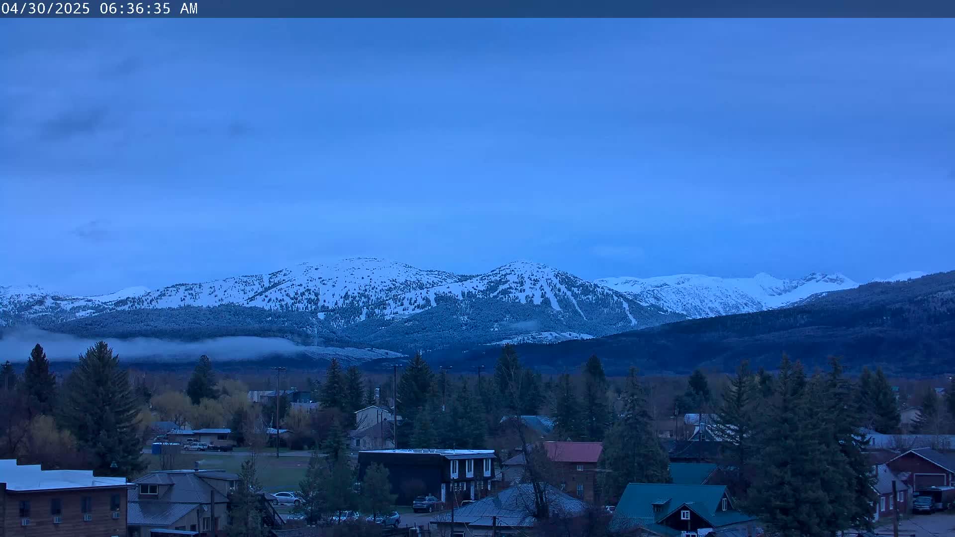 A small town sits before a range of snow-capped mountains under a twilight sky.