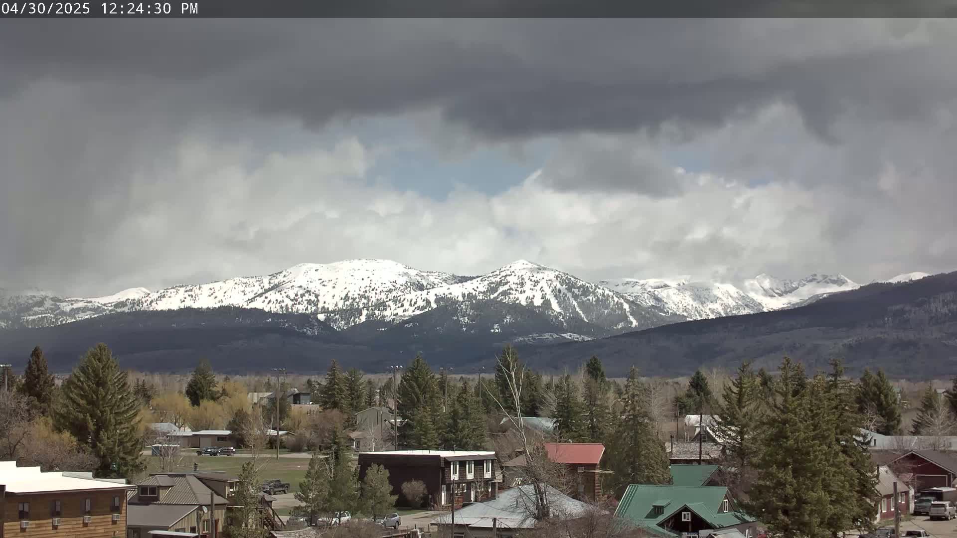 A small town with houses and evergreen trees sits in the foreground, under a partly cloudy sky, with snow-capped mountains in the background.