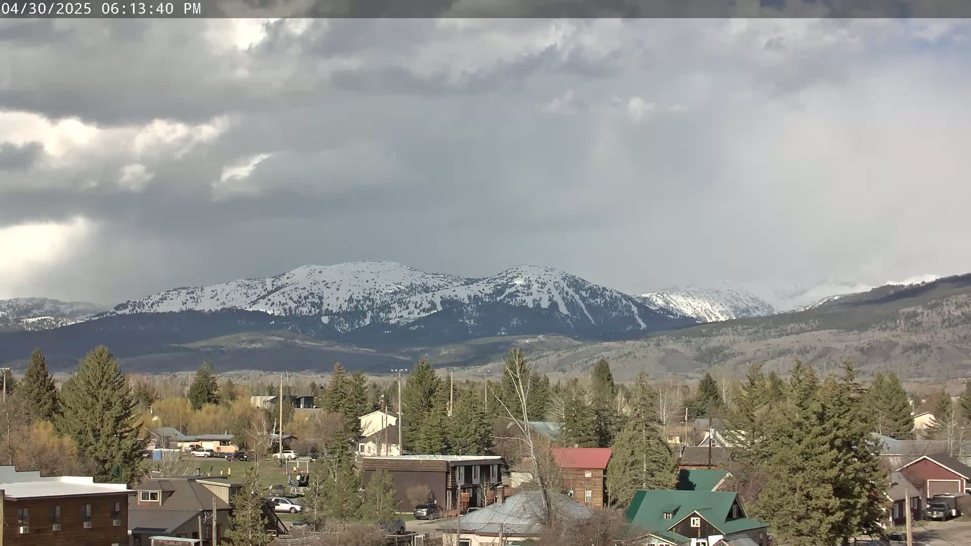 A small town nestled among evergreens sits before snow-capped mountains under a mostly cloudy sky.