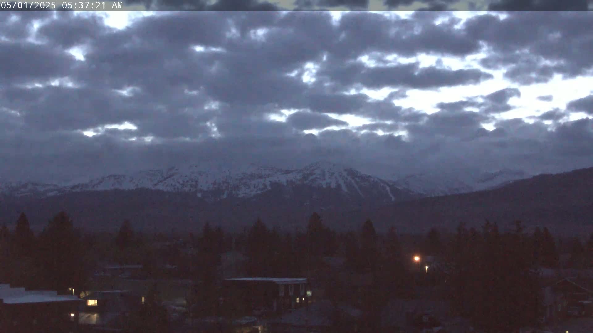 Snow-capped mountains are visible in the distance under a cloudy, pre-dawn sky above a dark, tree-lined town.