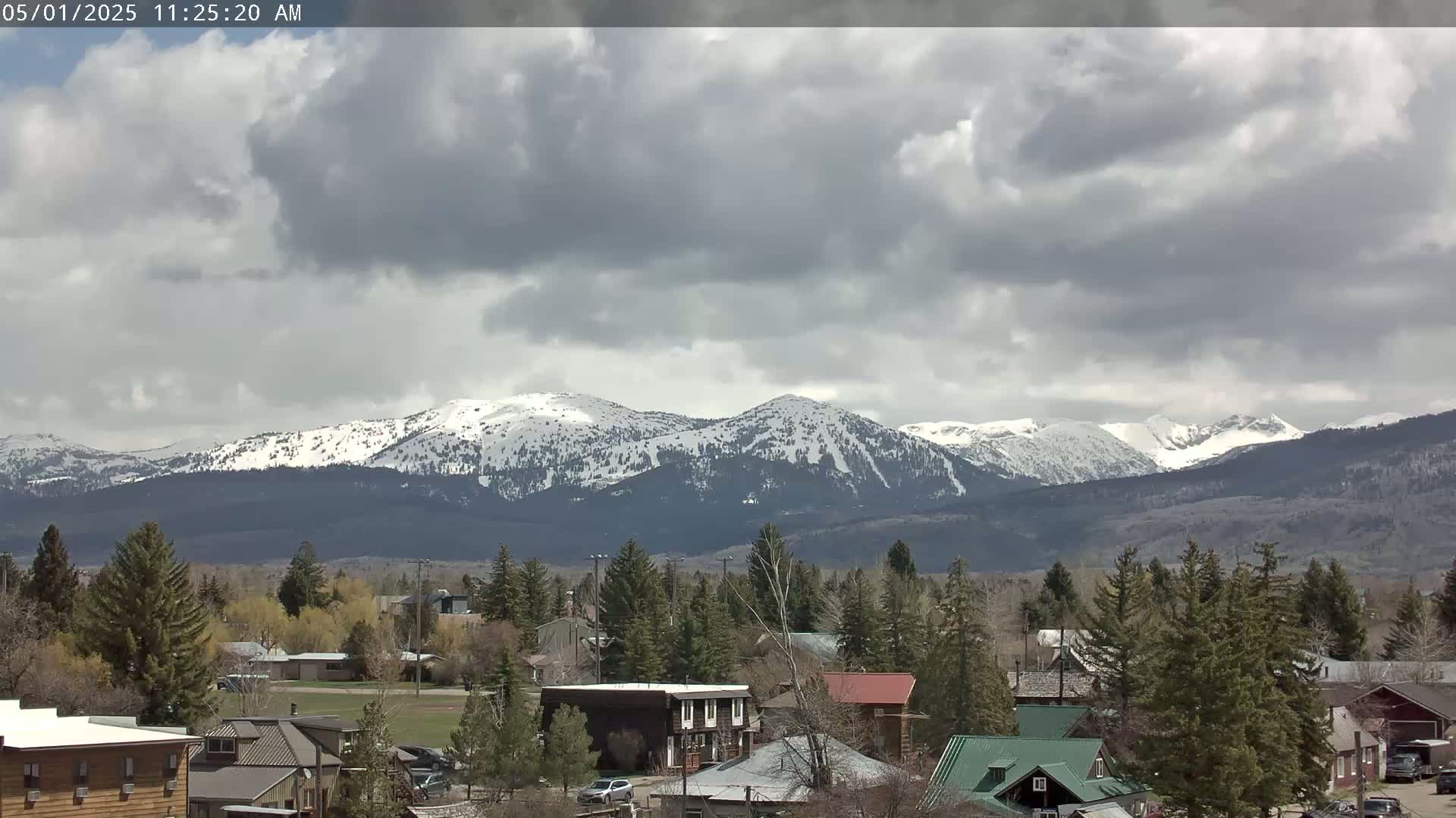 A small town nestled among evergreens, with snow-capped mountains in the background under a cloudy sky.