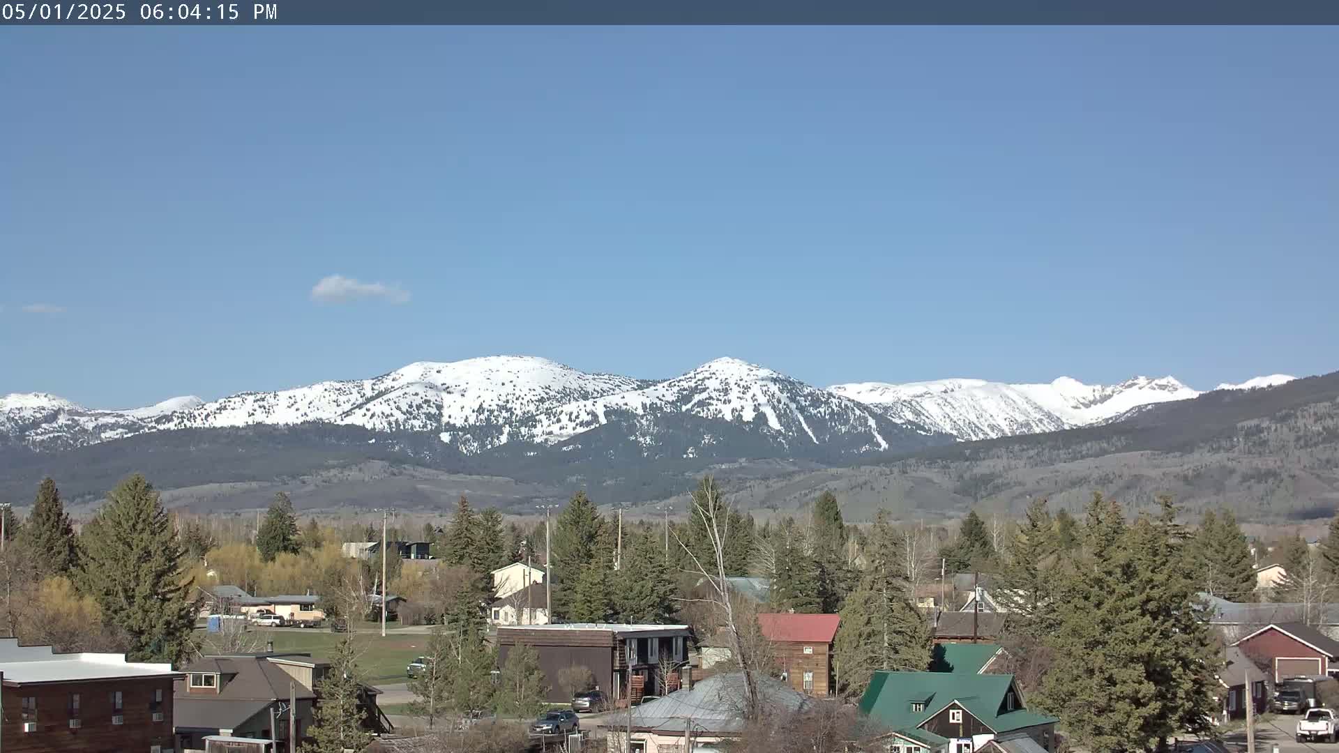 A small town nestled among evergreen trees sits before a snow-capped mountain range under a clear blue sky.