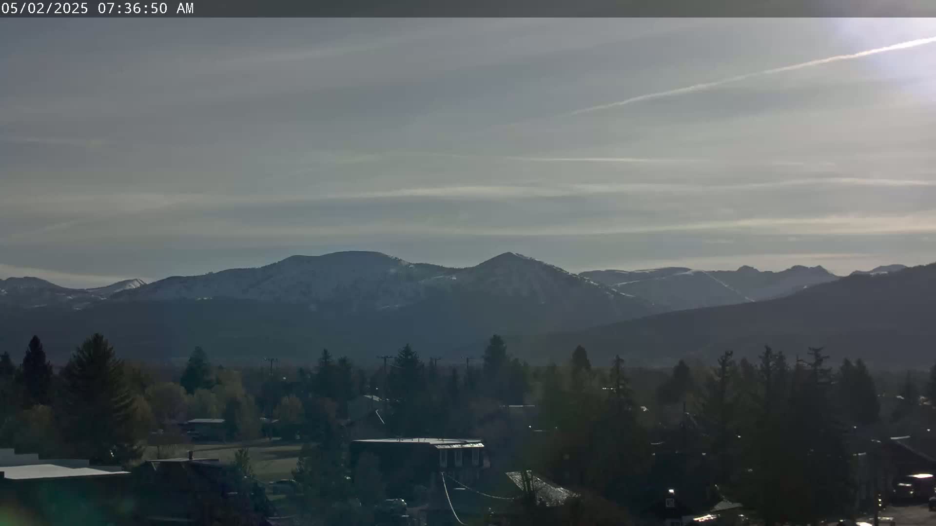 A snow-dusted mountain range is visible in the distance under a partly cloudy sky, behind a town of dark evergreen trees and buildings.