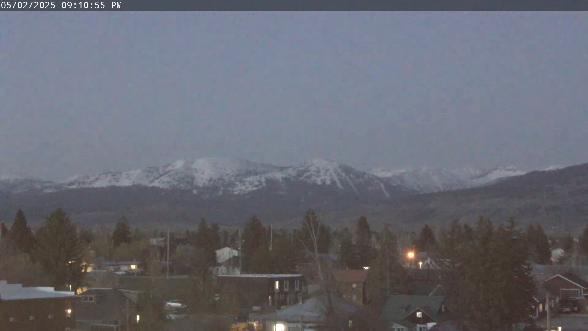 A town with houses and trees sits in the foreground, with snow-capped mountains visible in the distance under a dusky sky.
