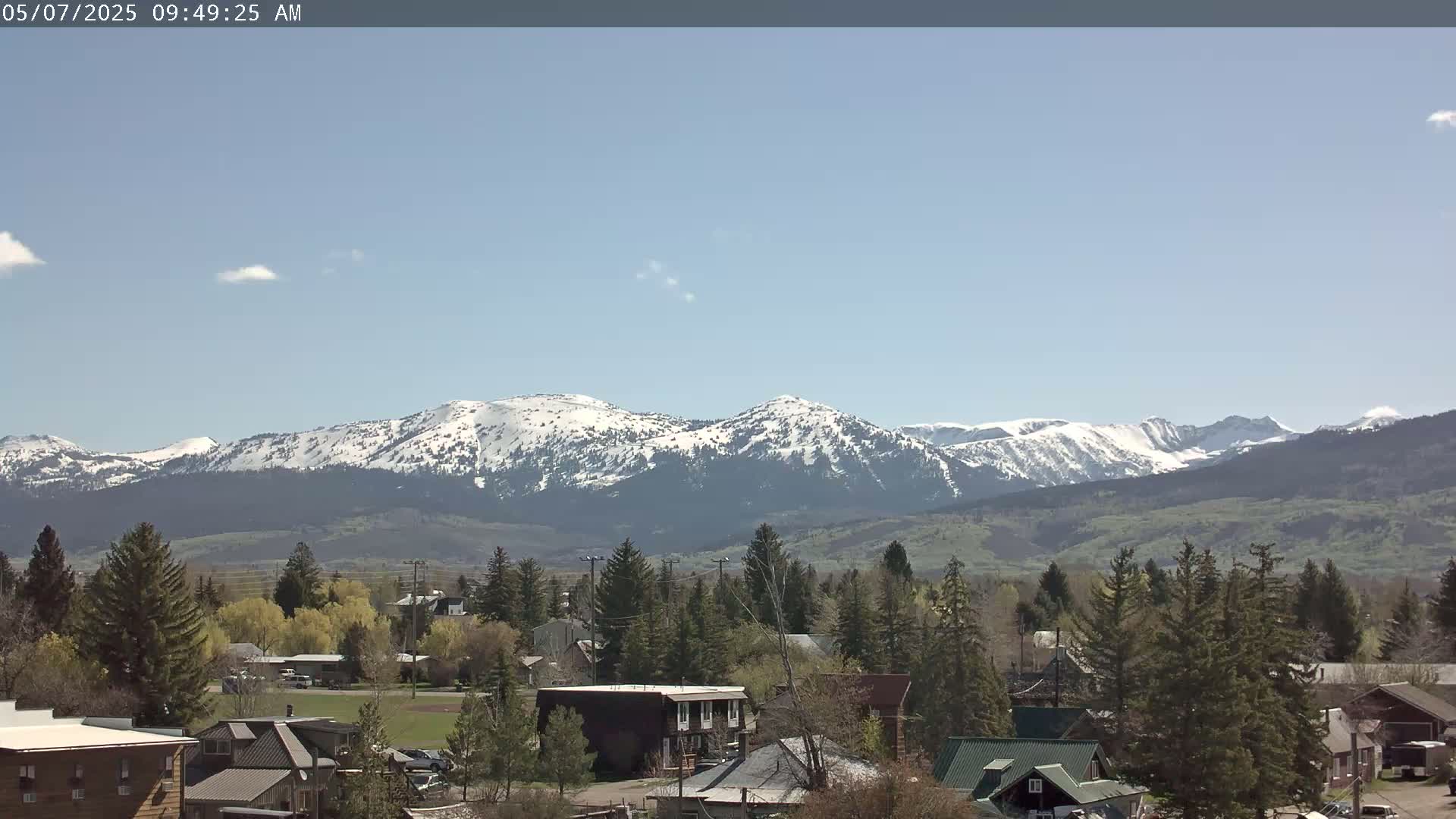 A small town with evergreen trees and houses sits in a valley, with snow-capped mountains visible in the background under a mostly clear, light blue sky.