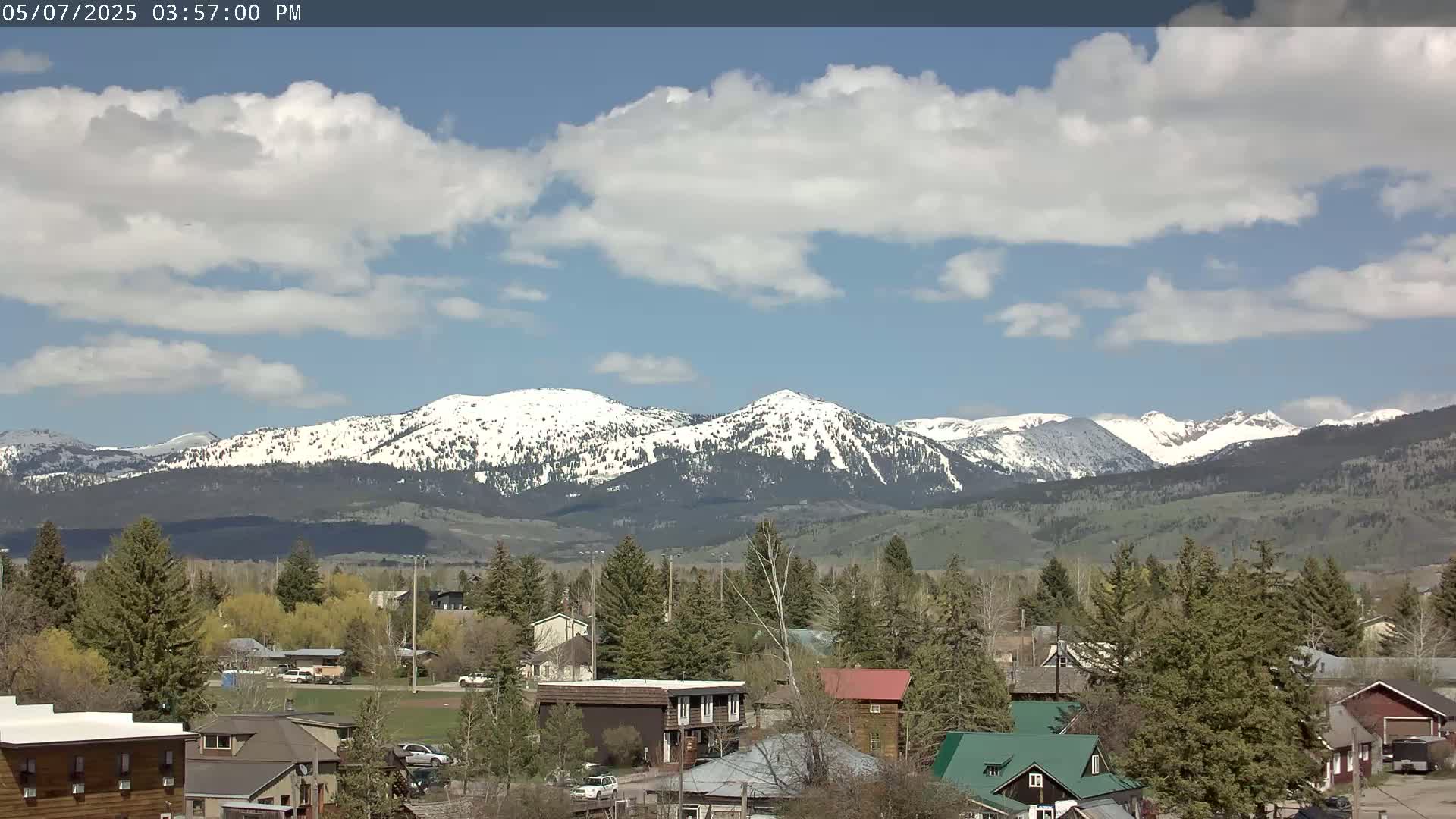 A partly cloudy day reveals a snow-capped mountain range in the background, overlooking a town of houses and evergreen trees.