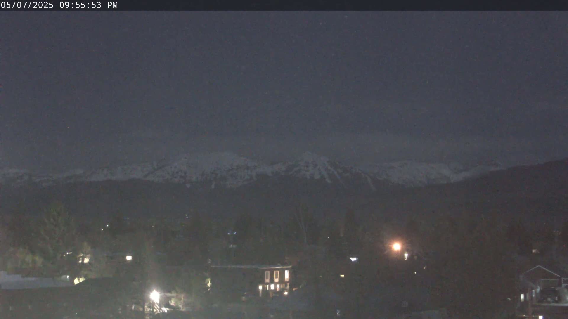 Snow-capped mountains are visible in the distance under a dark, lightly snowing night sky above a dimly lit town.