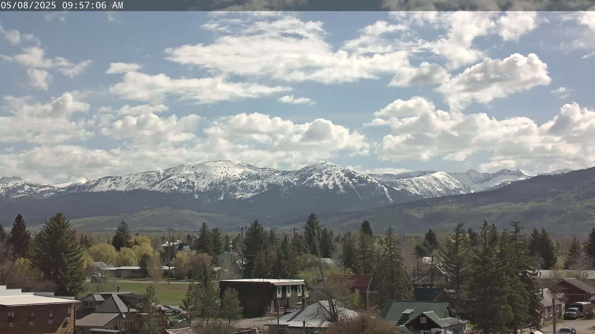A partly cloudy sky overlooks a small town nestled in a valley, with snow-capped mountains in the background.