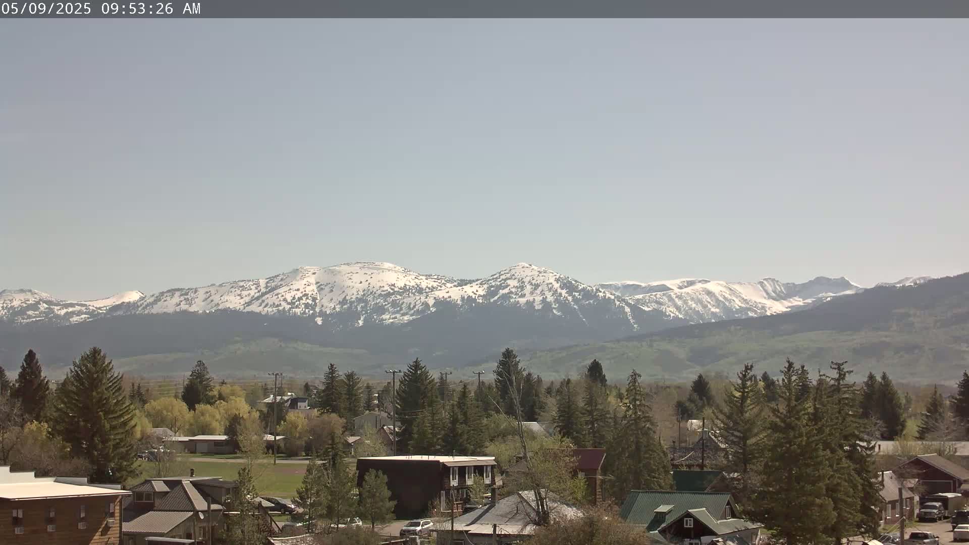 A small town nestled among evergreens, overlooks snow-capped mountains under a clear sky.