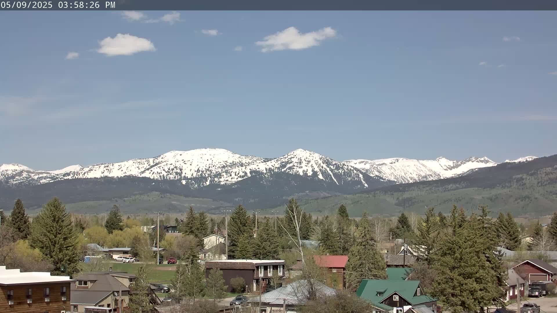 A small town nestled among green trees and shrubs, with snow-capped mountains in the background under a mostly clear, bright sky.