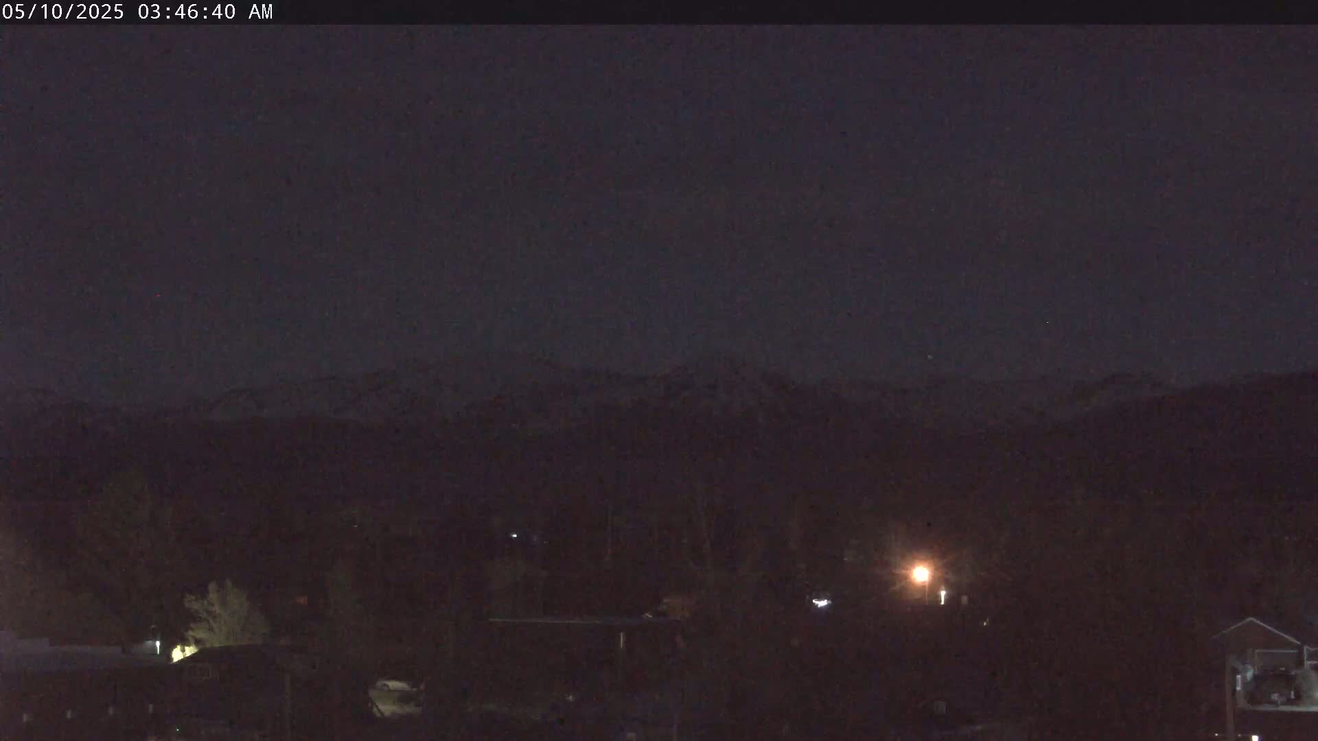 A nighttime view shows dark, snow-capped mountains in the distance, above a dimly lit town with scattered trees and buildings.