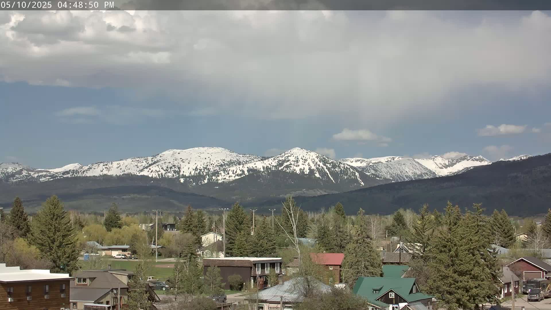 A partly cloudy day reveals snow-capped mountains in the distance behind a small town with houses and evergreens.