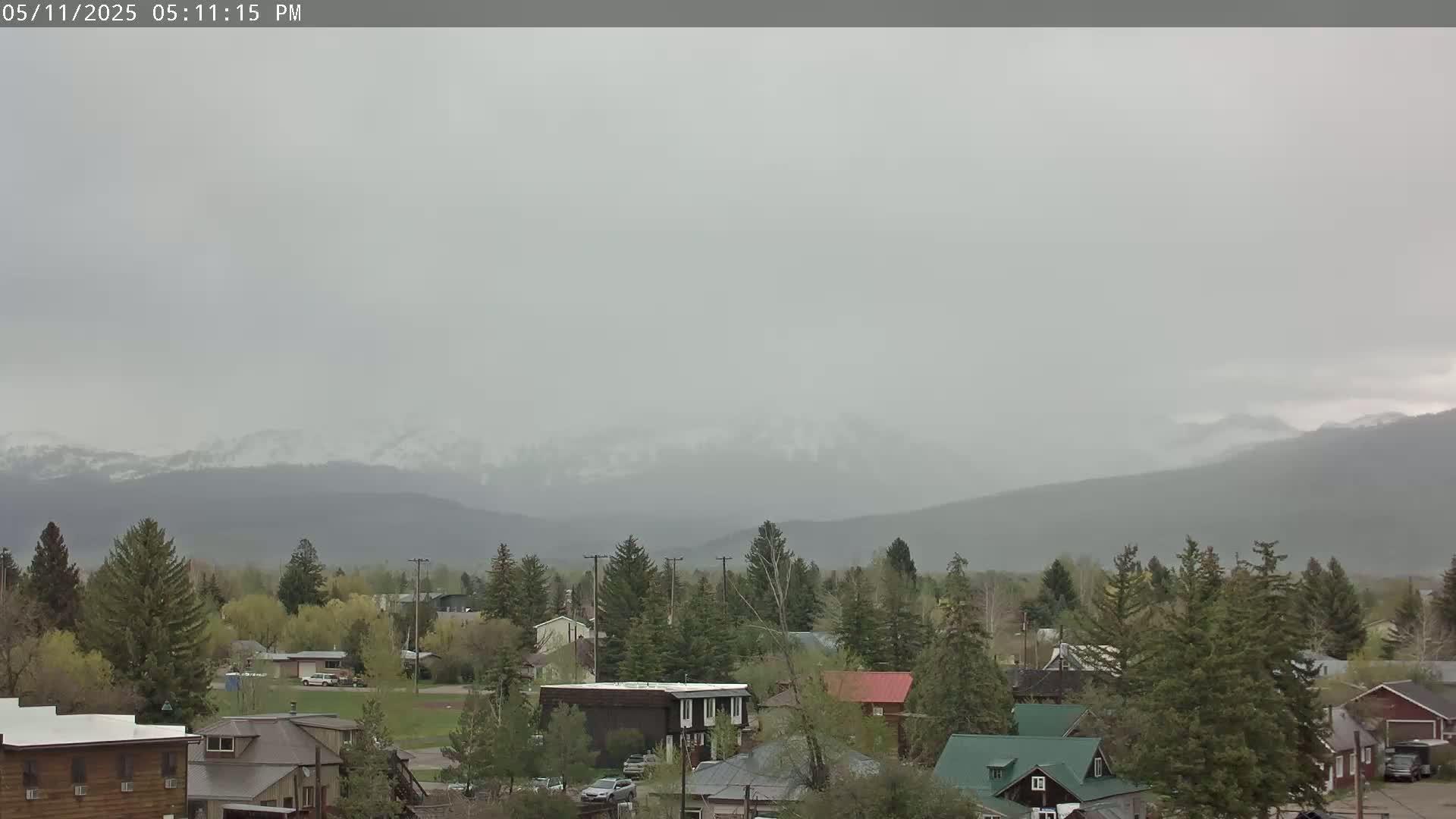 A small town with houses and evergreen trees sits in the foreground, with hazy mountains showing snow-capped peaks in the background under an overcast sky.