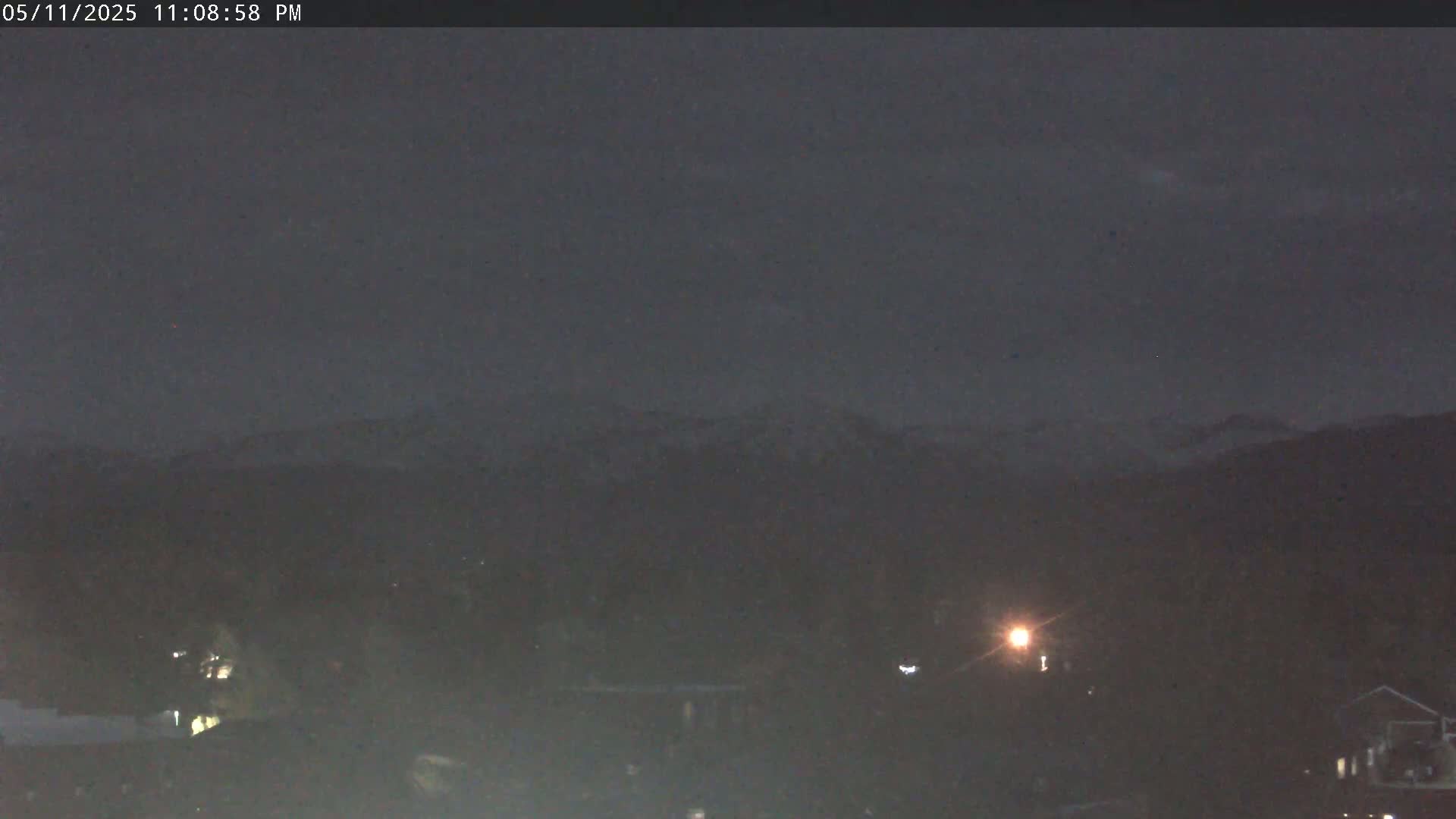 A nighttime long shot shows a dimly lit town with snow-capped mountains in the distance under a dark, overcast sky.