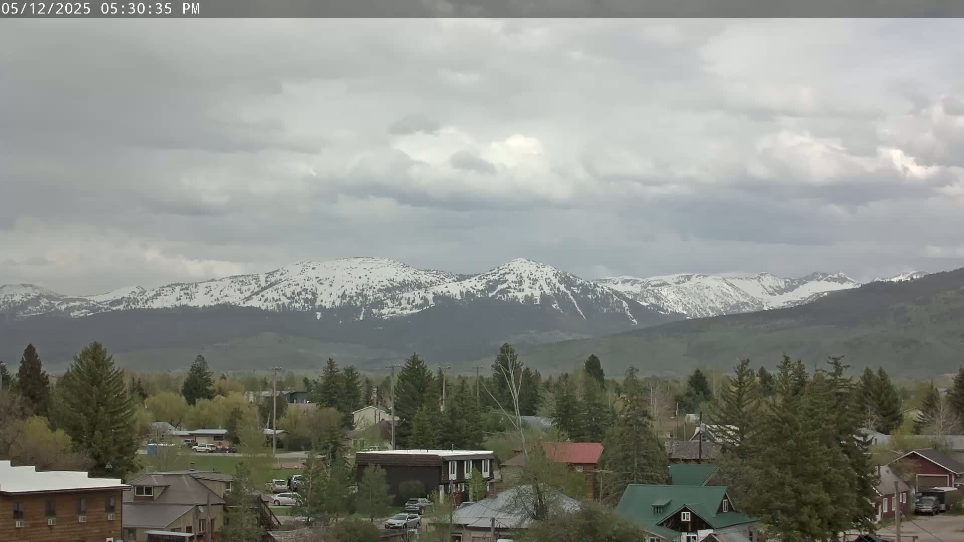 A small town nestled amongst evergreens sits before a snow-dusted mountain range under a cloudy sky.