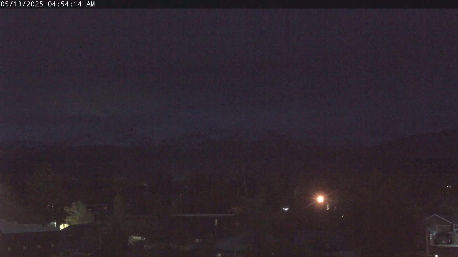 A dark nighttime scene shows a mostly obscured landscape with distant mountains under a clear, dark sky, illuminated by a few scattered lights.