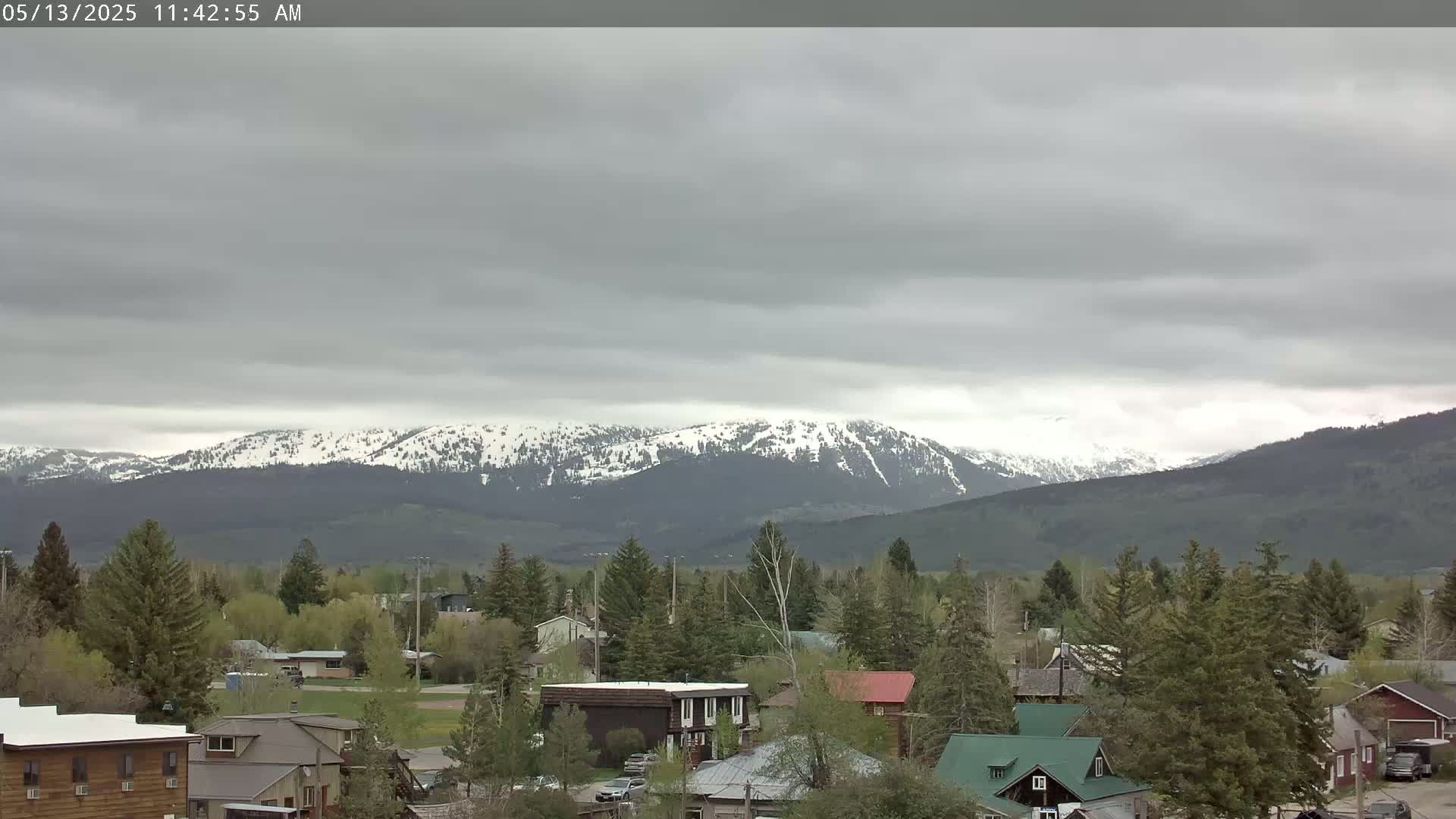 A small town nestled among evergreen trees, with snow-capped mountains in the background under an overcast sky.