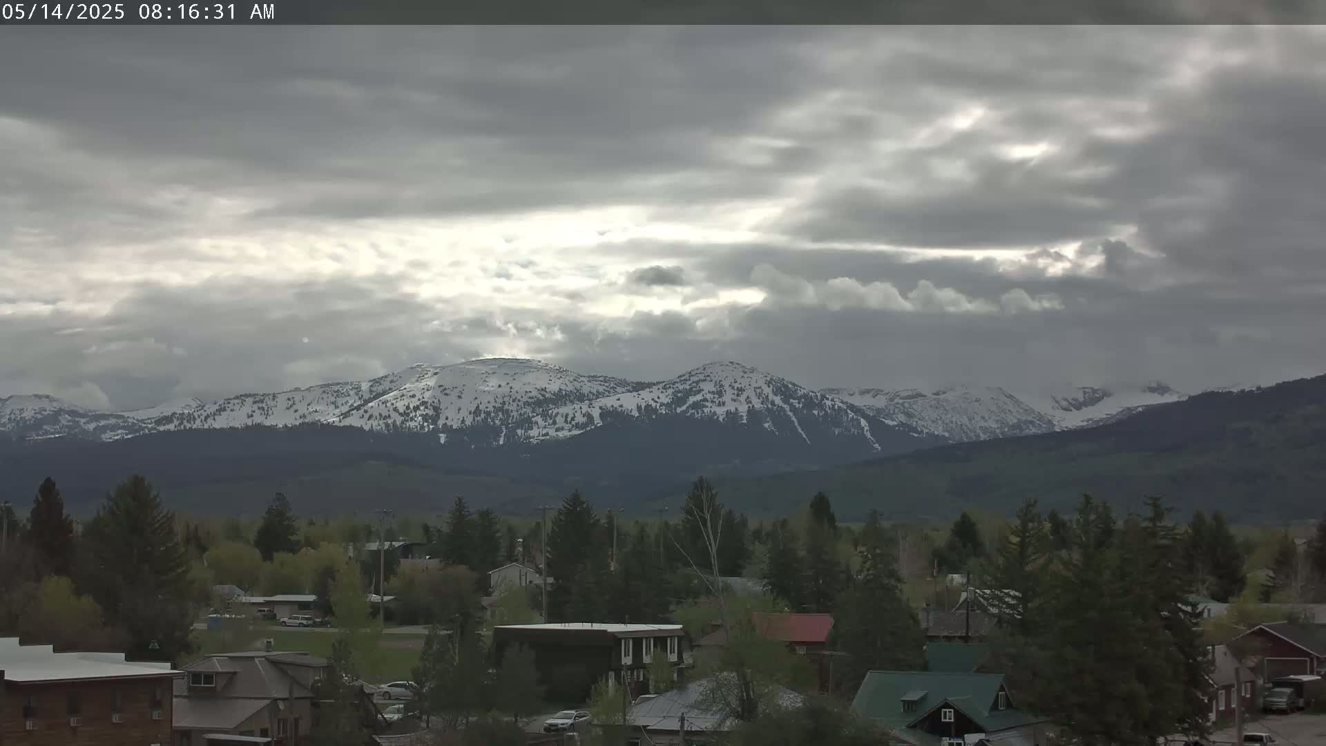 A small town nestled among green trees and snow-capped mountains under a mostly cloudy sky.