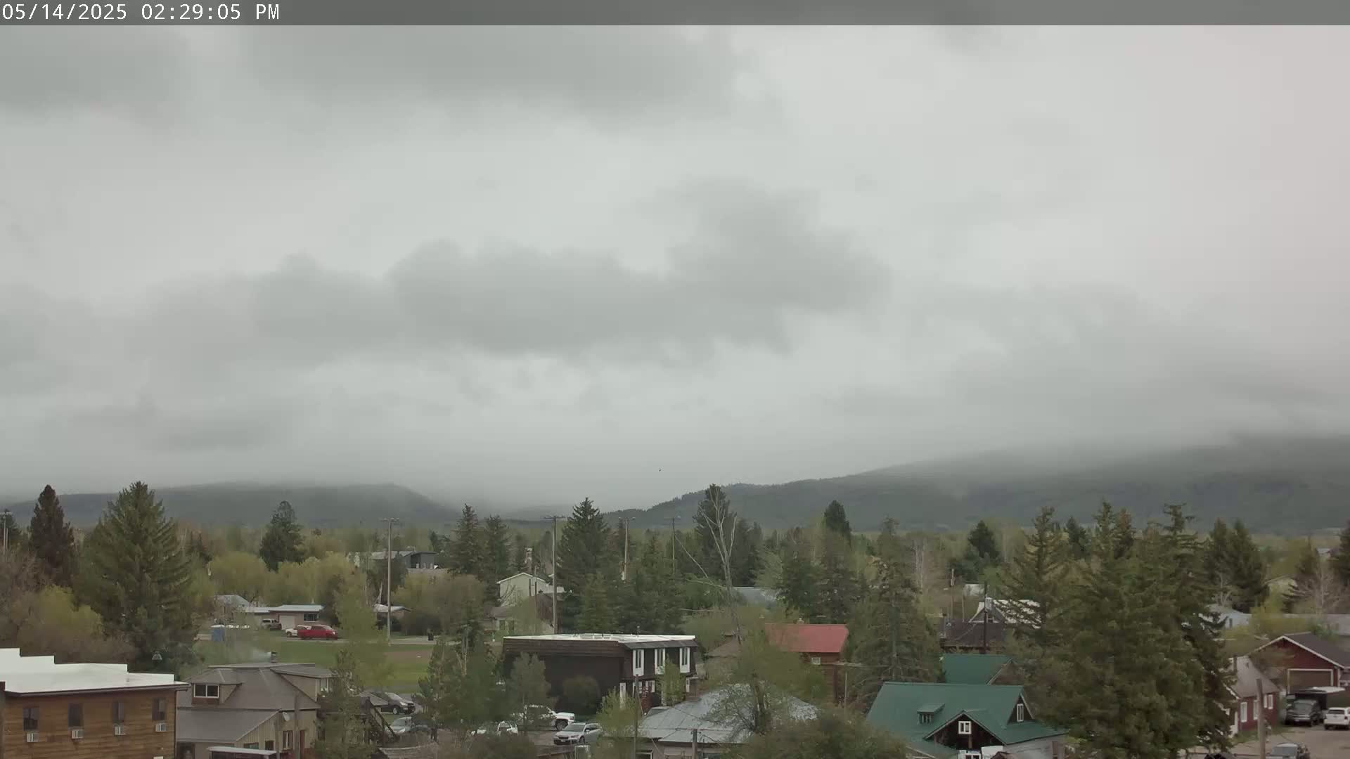 A small town nestled among evergreen trees sits beneath a heavily overcast sky, with hazy mountains visible in the distance.