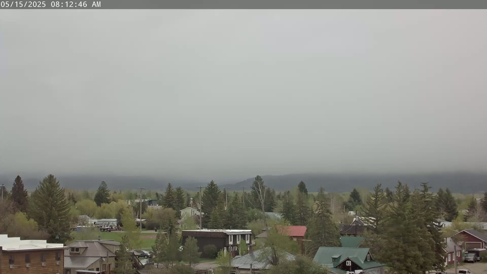 A hazy, overcast sky hangs over a small town nestled amongst evergreen trees and distant, fog-shrouded mountains.