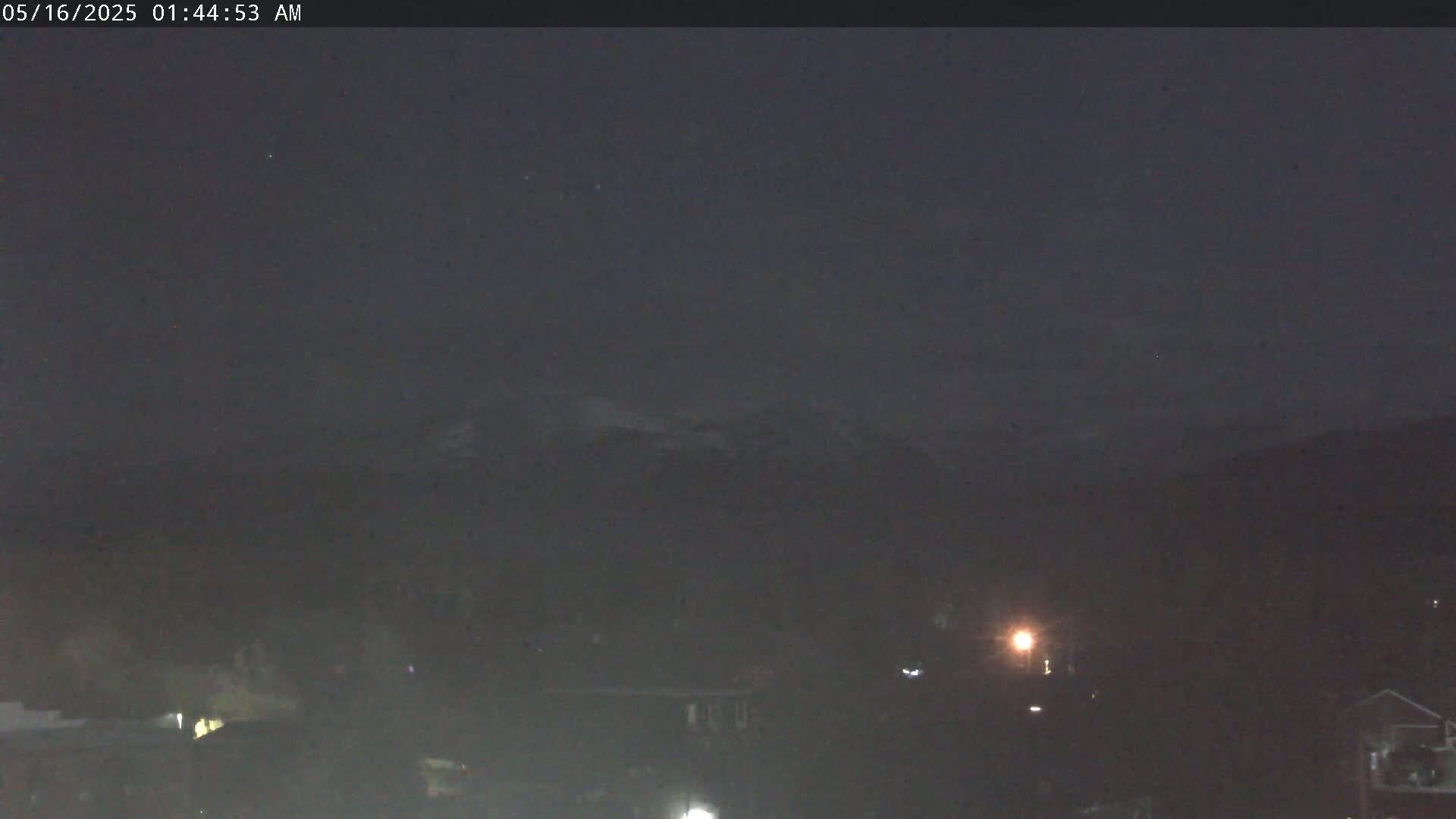 A dark, nighttime scene shows a distant mountain range under a mostly clear sky, with a few dim lights visible in the foreground.