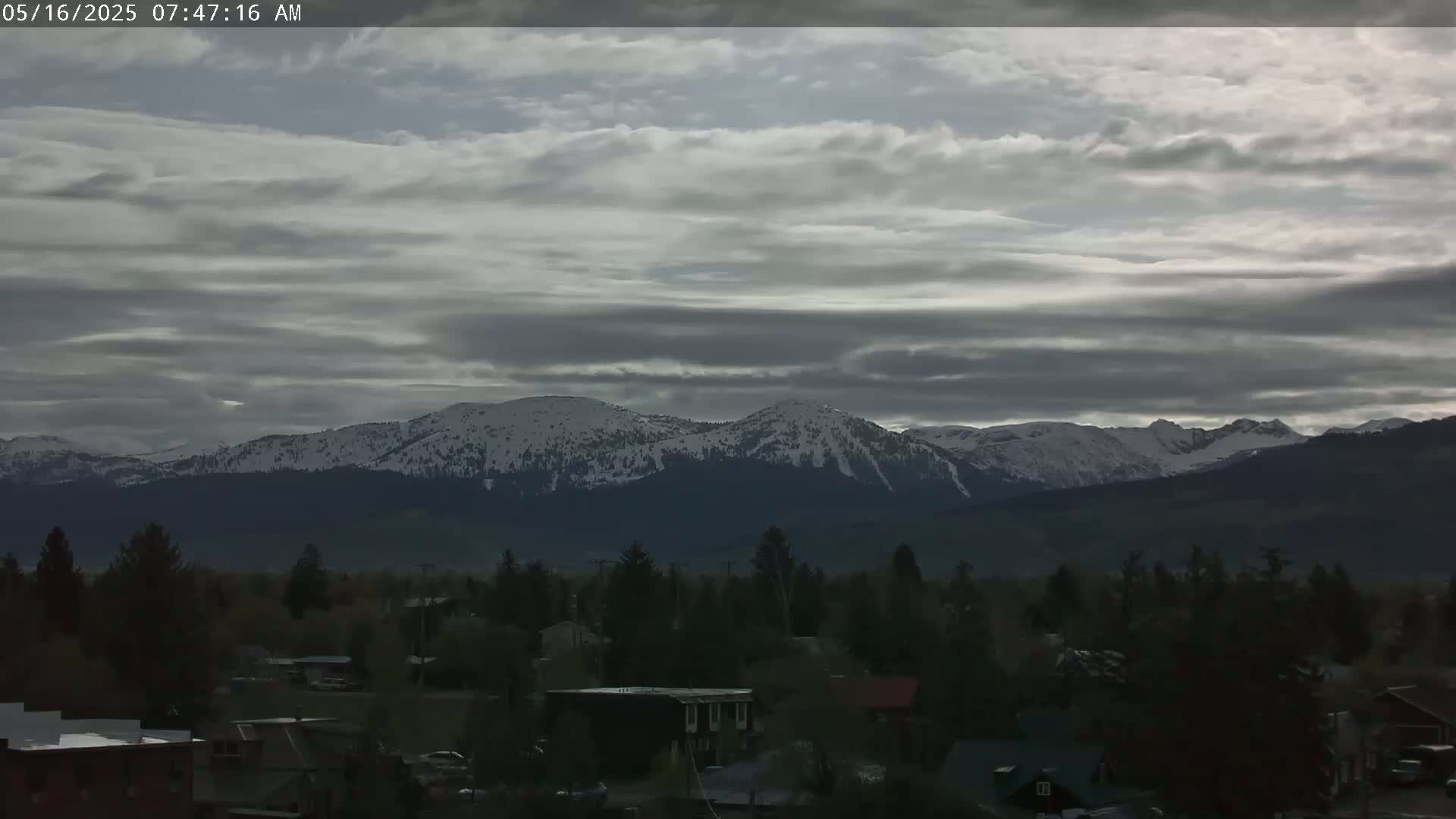 A snow-capped mountain range is visible in the distance under a cloudy sky, behind a town with houses and trees.