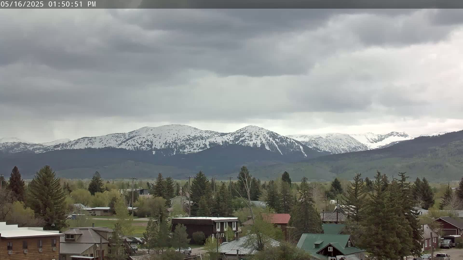 A small town nestled among evergreens overlooks snow-capped mountains under an overcast sky.