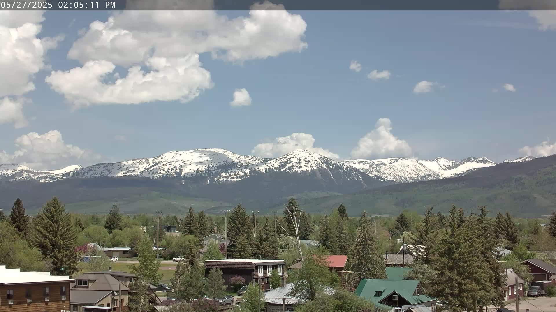A partly cloudy day reveals a snow-capped mountain range behind a small town nestled amongst green trees.