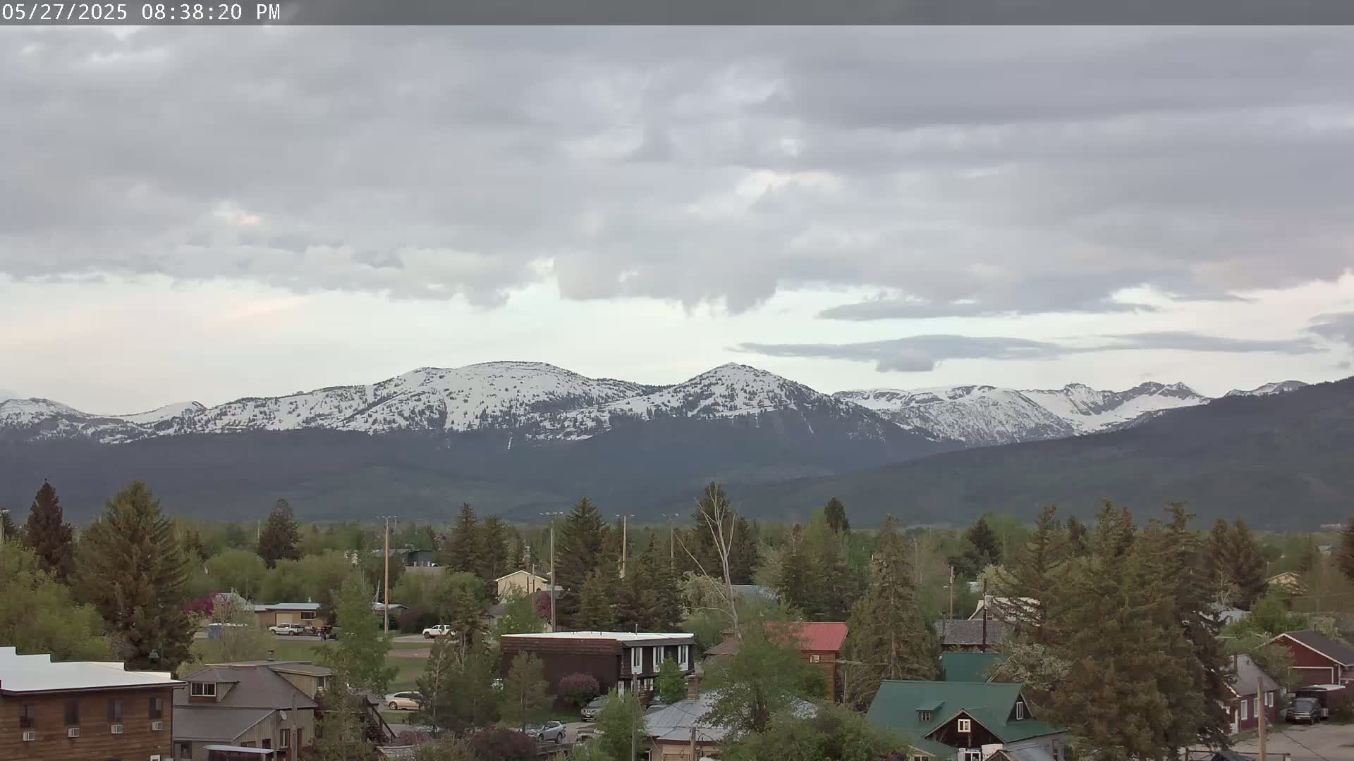 A small town with houses and trees sits in the foreground, under a cloudy sky, with snow-capped mountains in the background.