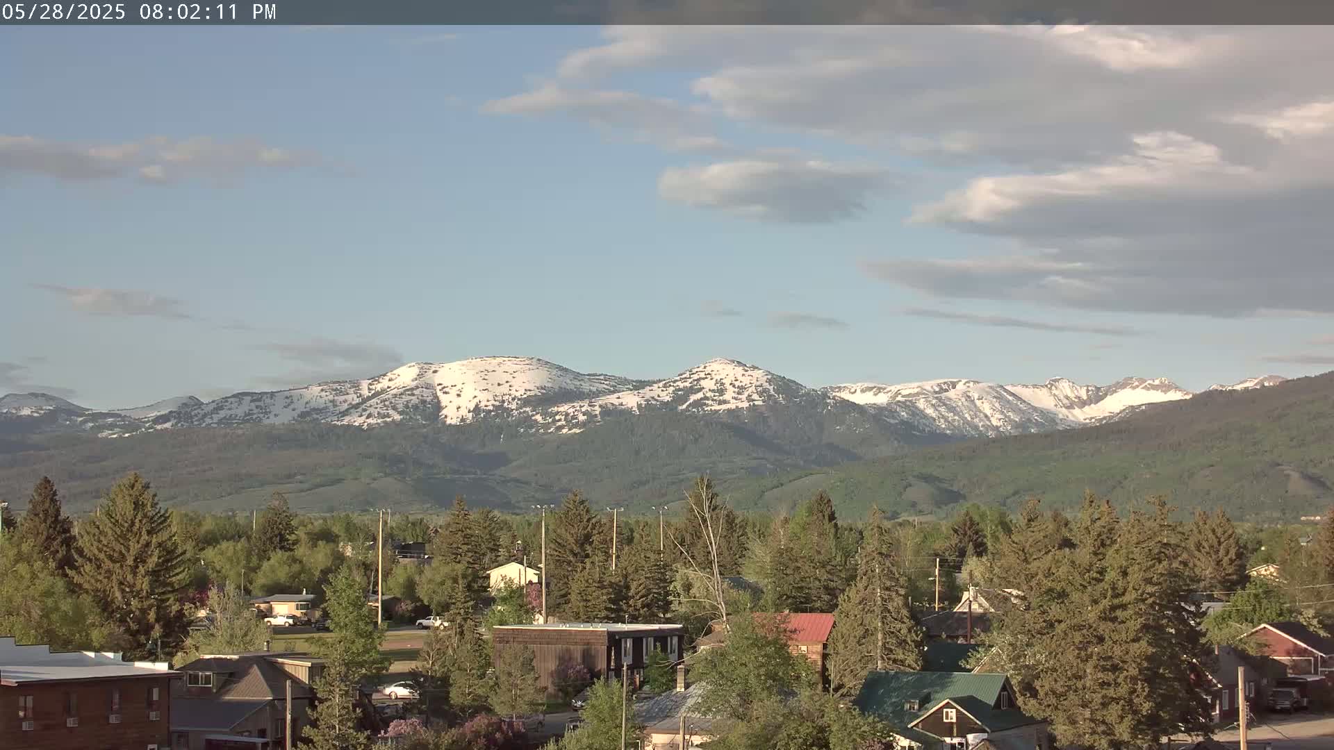 A partly cloudy afternoon view of a small town nestled among evergreens, with snow-capped mountains in the background.