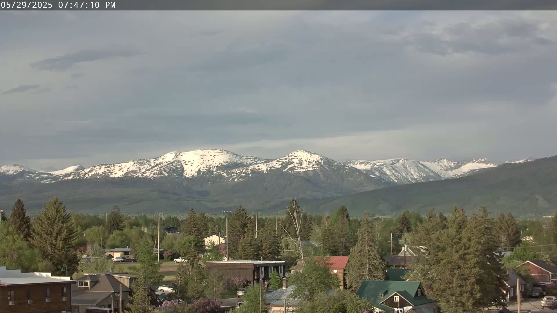 A small town nestled among evergreen trees, overlooks a mountain range with snow-dusted peaks under a mostly cloudy sky.