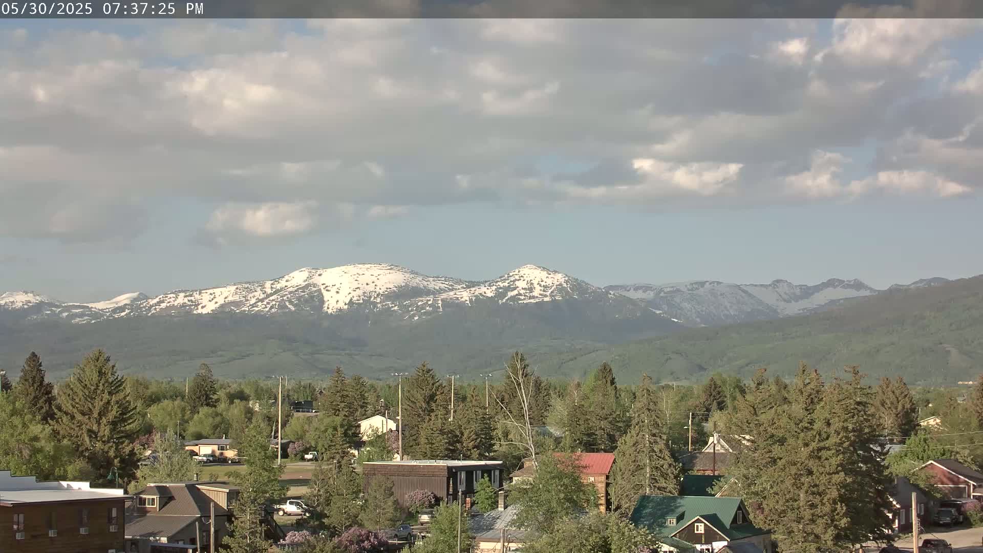 A partly cloudy sky overlooks a town nestled among green trees with snow-capped mountains in the background.