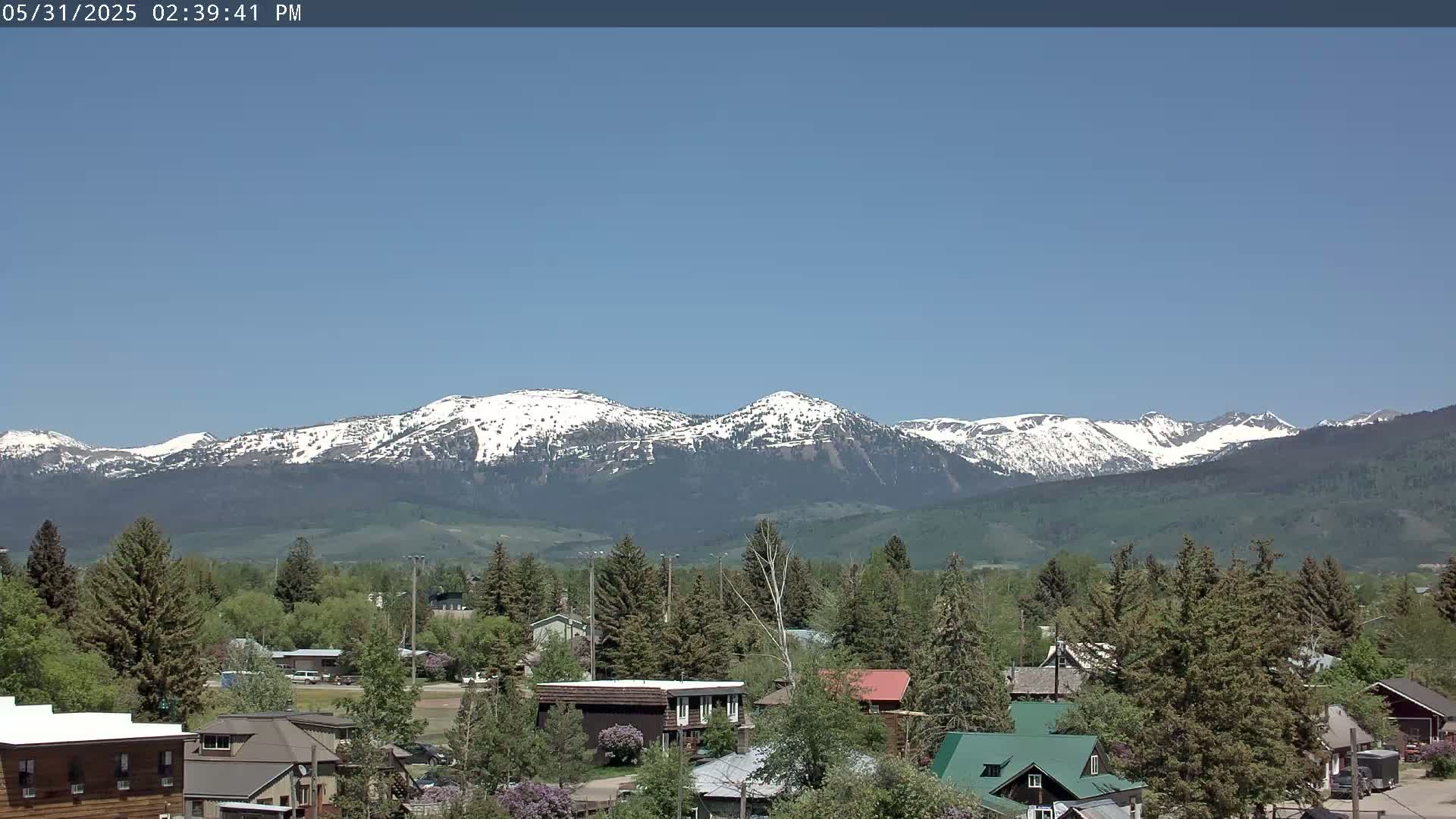 A small town nestled among evergreen trees sits before a mountain range with snow-capped peaks under a clear blue sky.