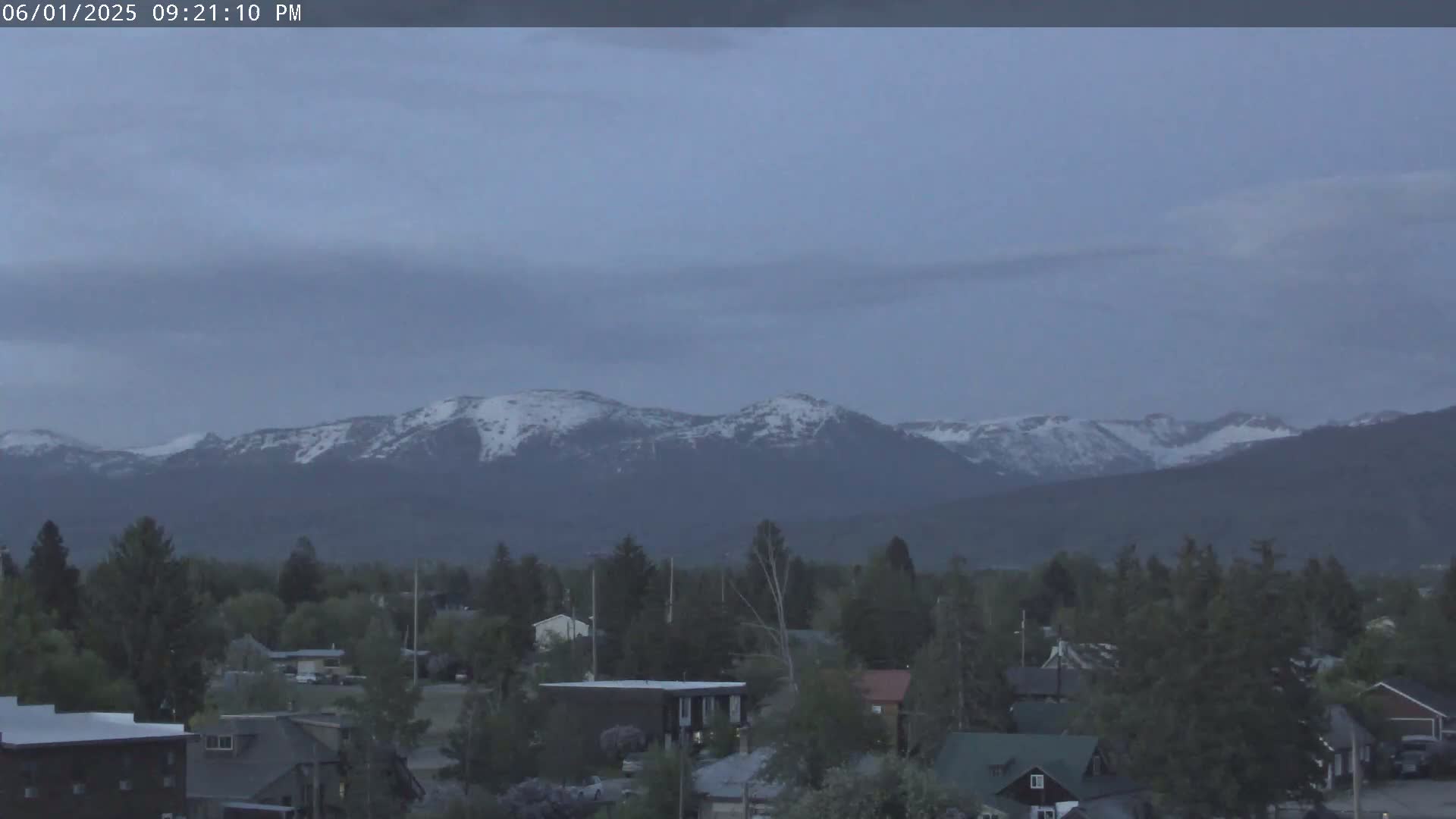 A small town sits before a range of snow-capped mountains under a dusky, overcast sky.
