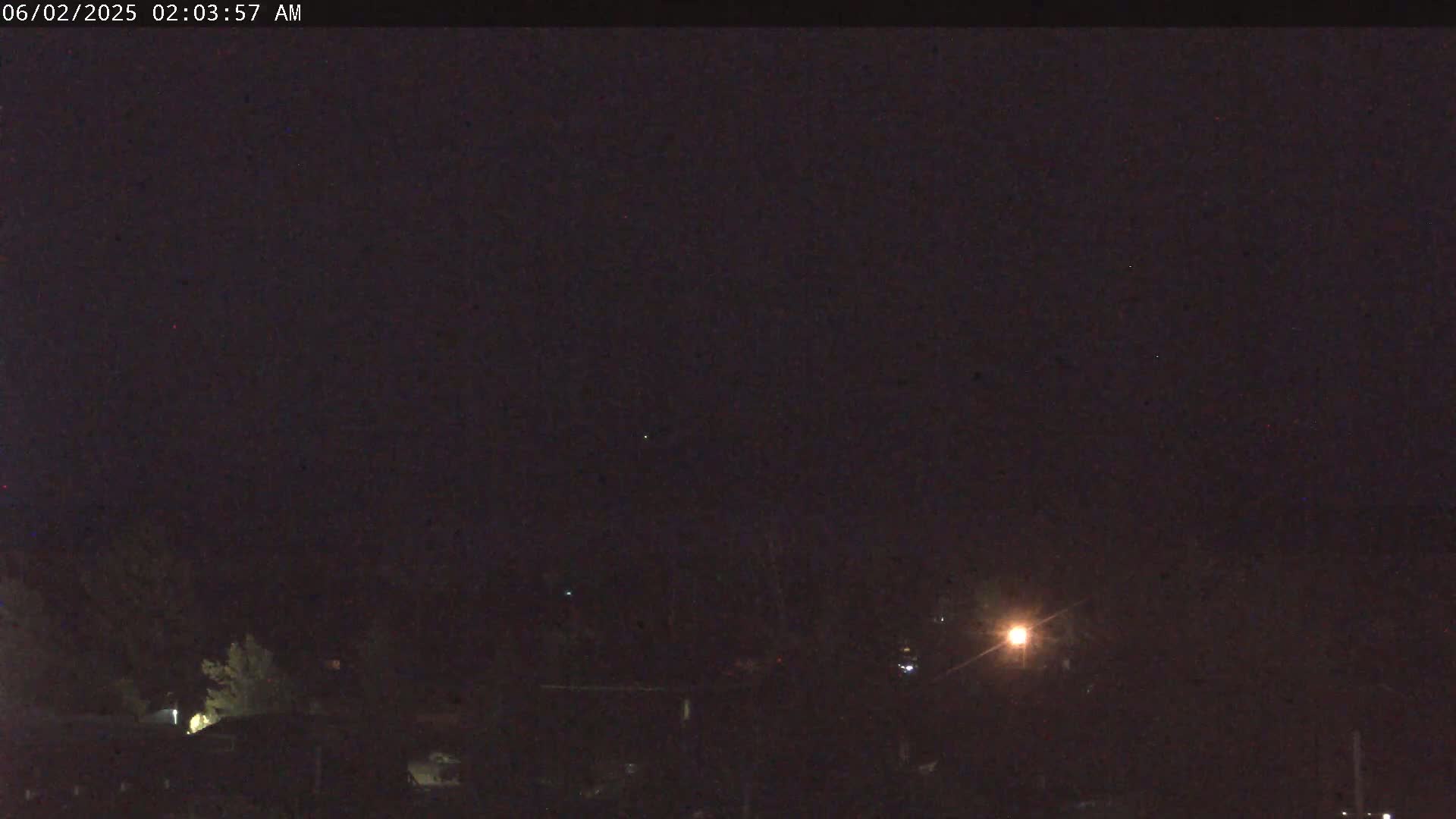 A nighttime, dark image shows a few dimly lit buildings and trees under a clear, dark sky.