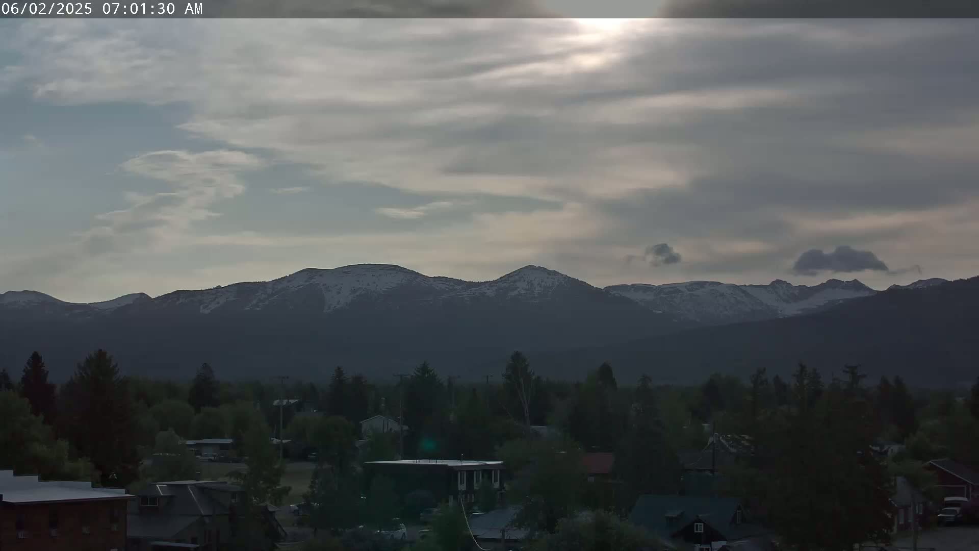A small town sits in a valley, nestled beneath a range of snow-capped mountains under a mostly cloudy, dusky sky.