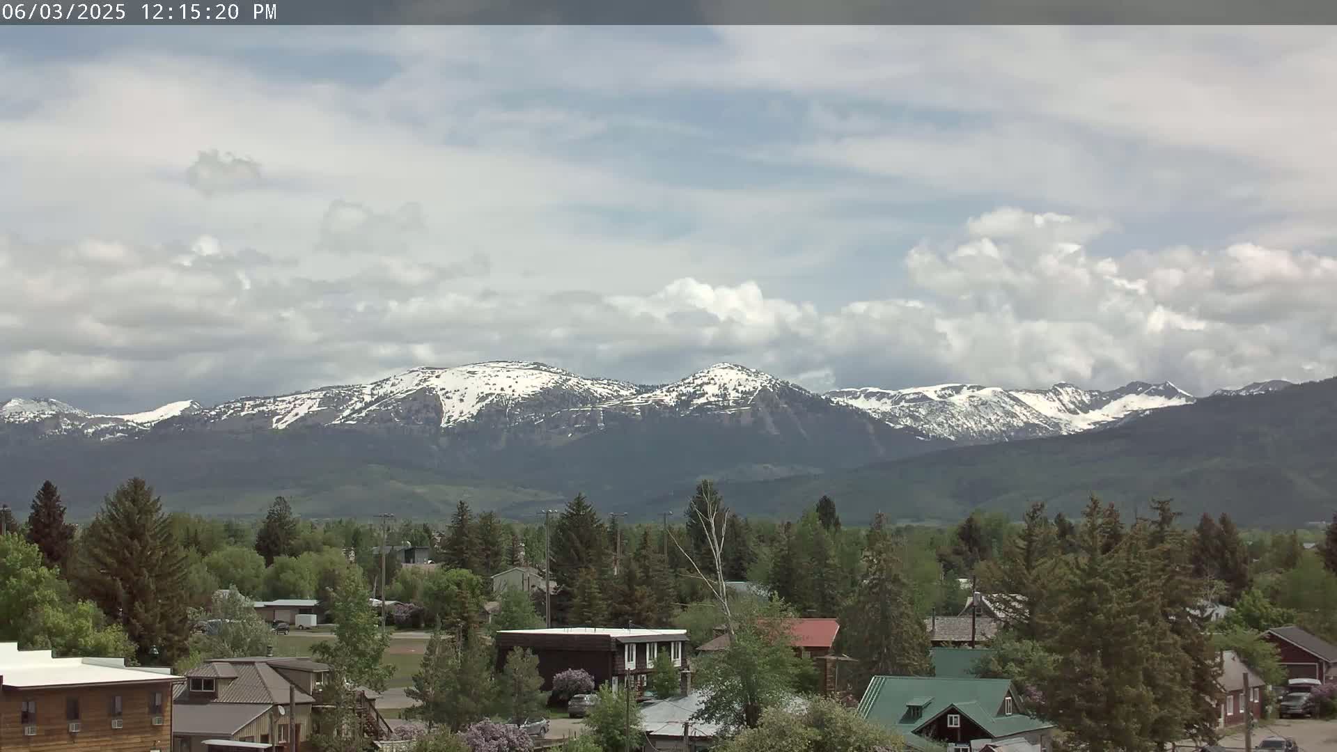 A partly cloudy day reveals a small town nestled amongst green trees, with snow-capped mountains in the background.