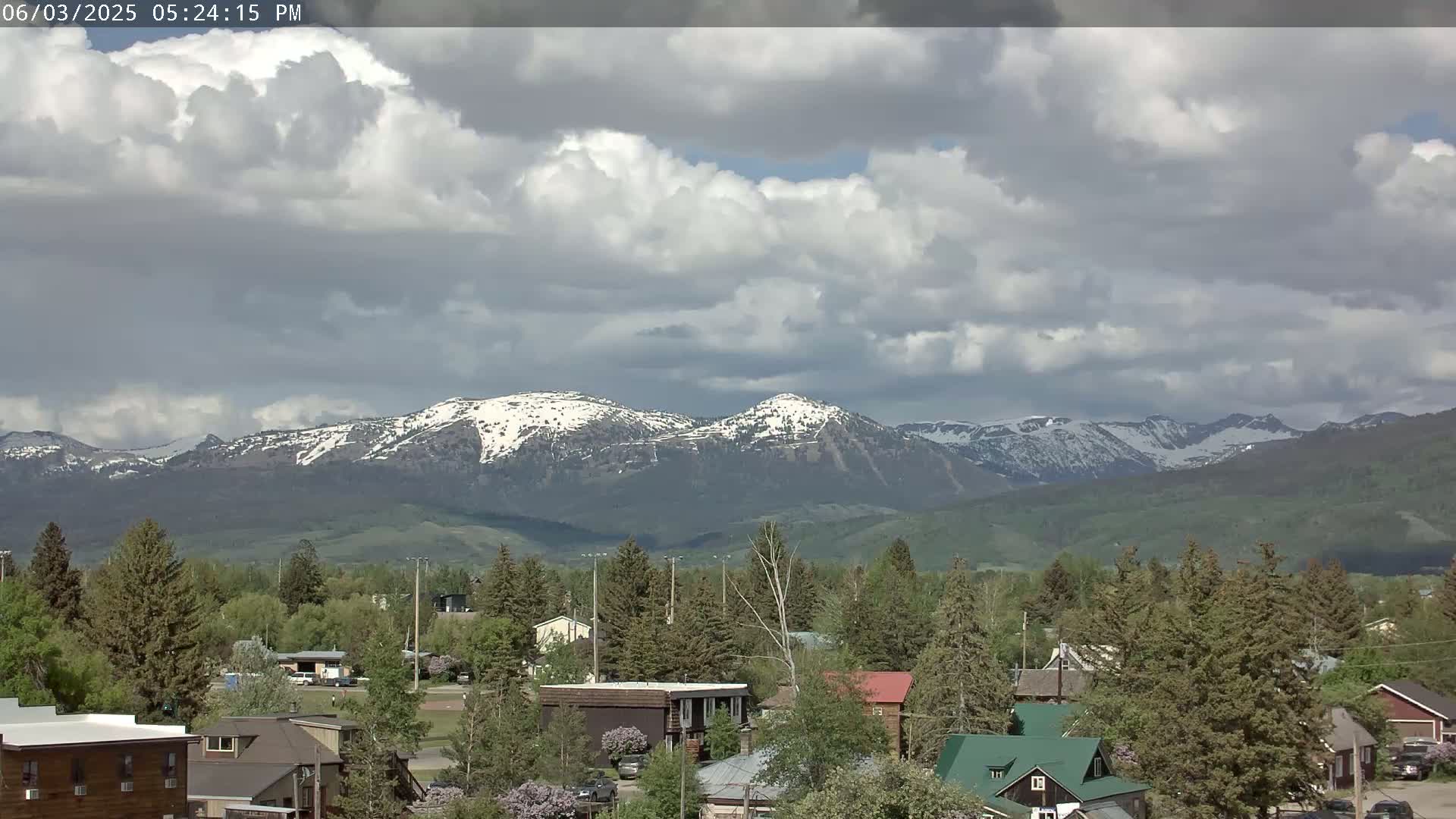 A partly cloudy day reveals snow-capped mountains in the distance, overlooking a town with houses and evergreen trees.
