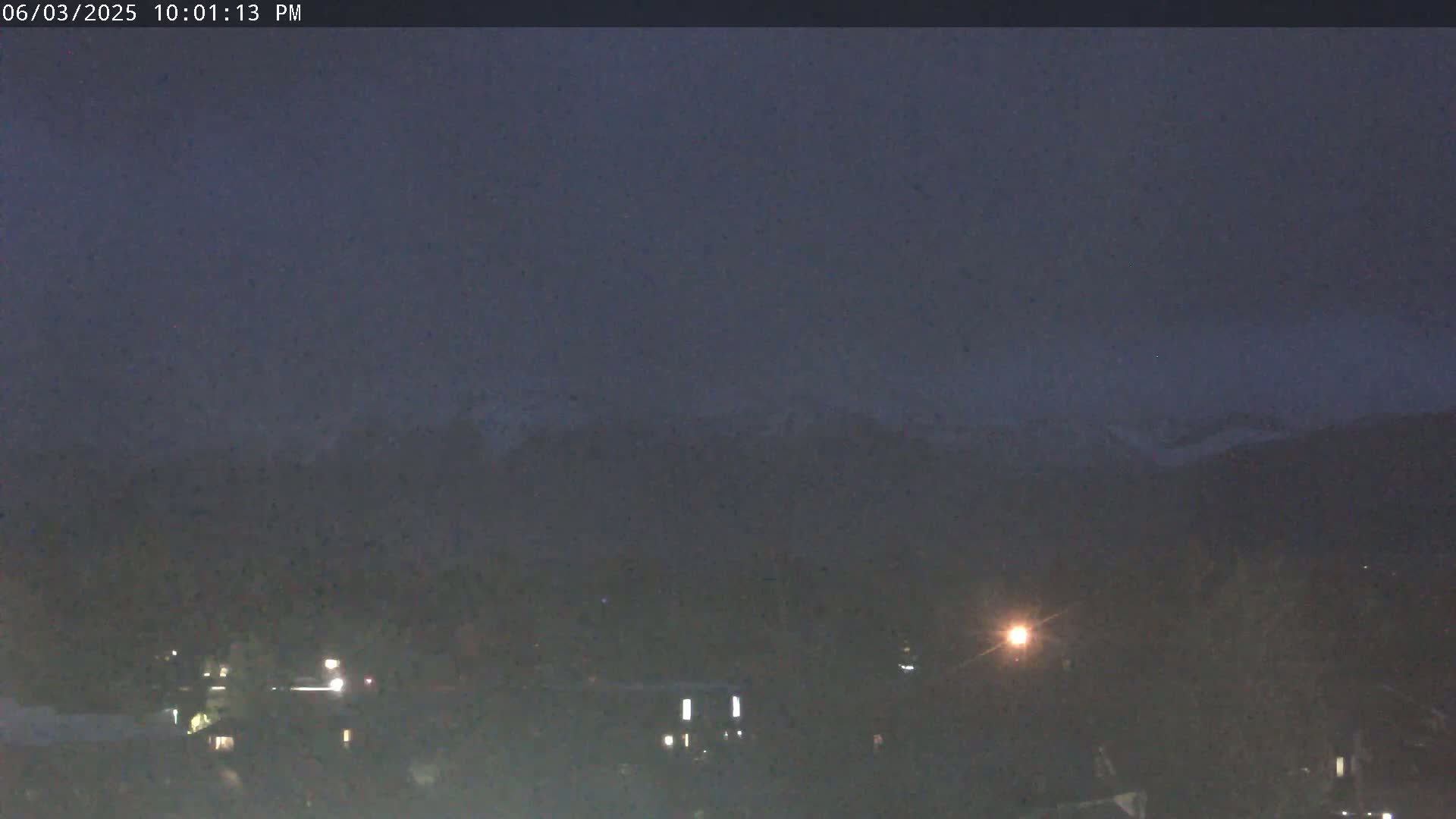 A nighttime view of snow-capped mountains in the distance, over a dimly lit town under a dark sky.