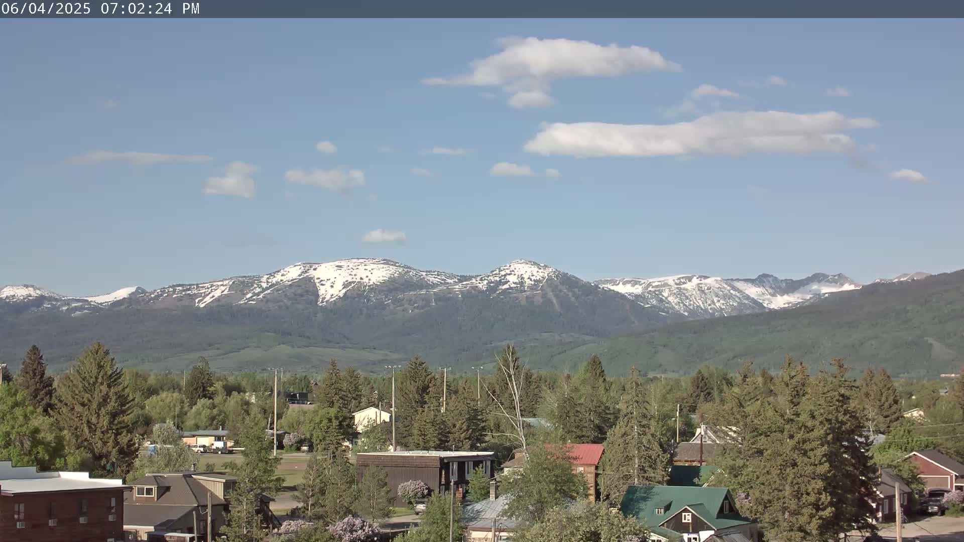 A small town nestled among green trees and shrubs, with snow-capped mountains in the background under a mostly clear, bright blue sky.