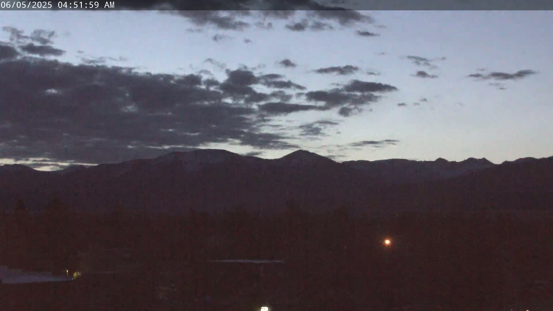 A dark, overcast pre-dawn sky illuminates snow-capped mountains in the distance, with a flat, dark foreground.