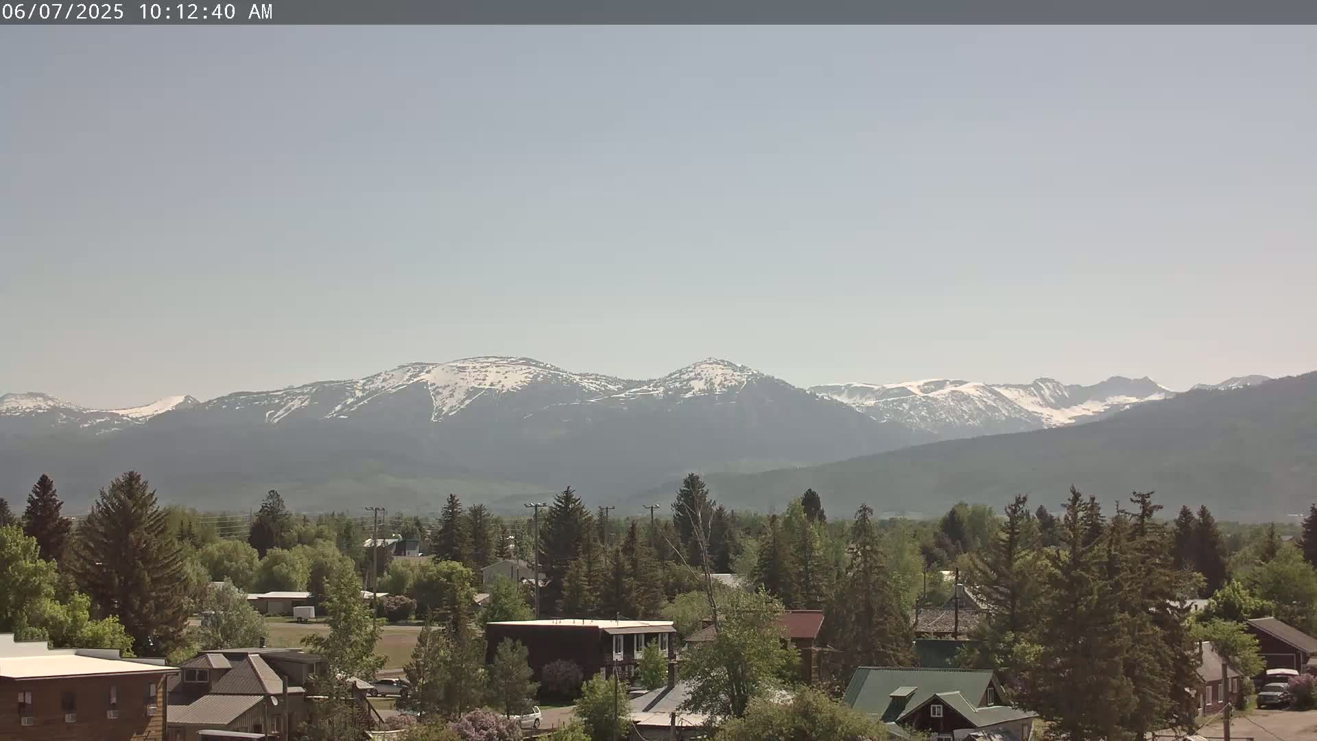 A hazy, sunny day reveals snow-capped mountains in the distance, overlooking a town nestled among evergreen trees.