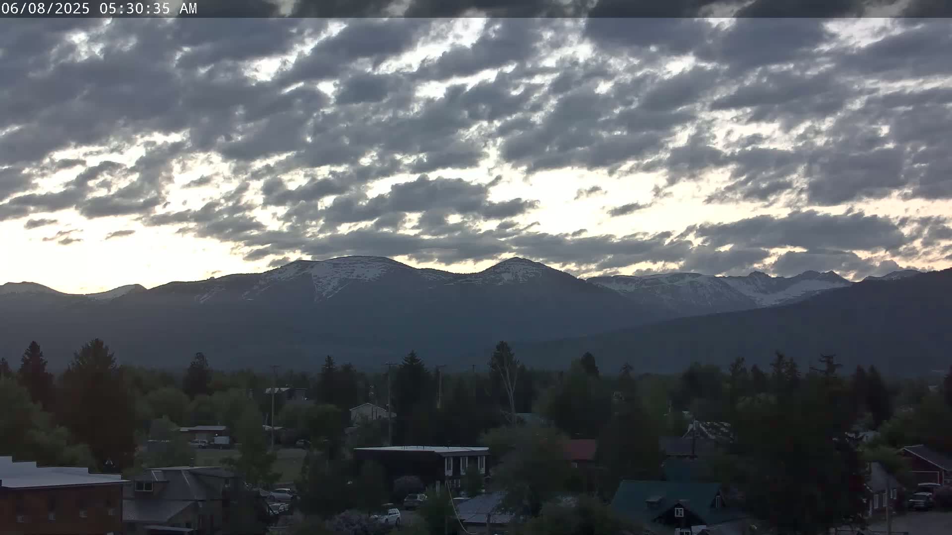 A partly cloudy sunrise illuminates snow-capped mountains behind a small town.