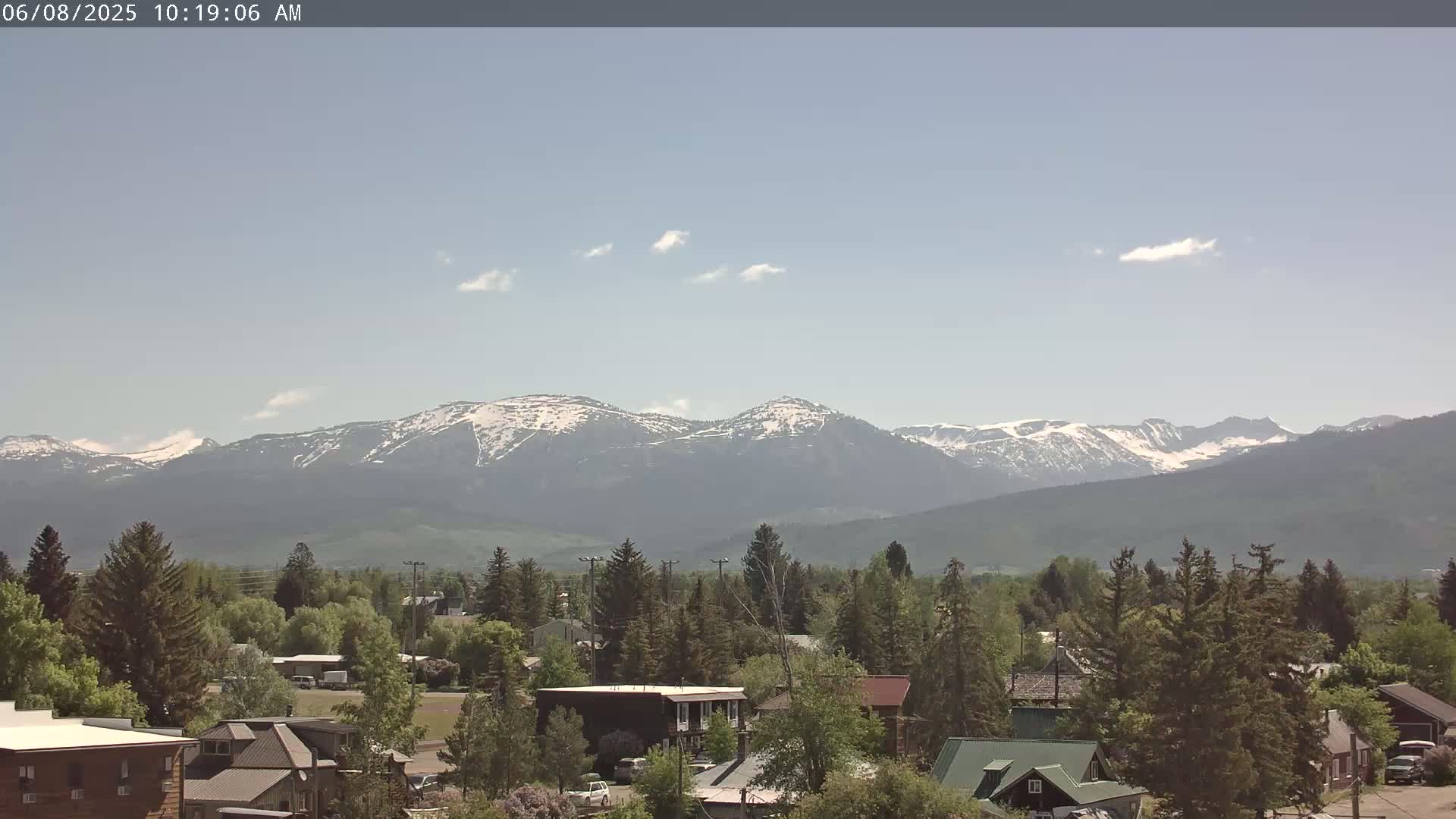 A small town nestled among evergreen trees overlooks a distant mountain range partially covered in snow under a mostly clear sky.