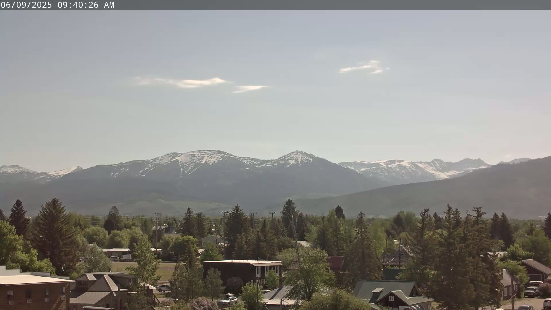 A small town nestled among evergreen trees, with snow-capped mountains in the background under a hazy, mostly clear sky.