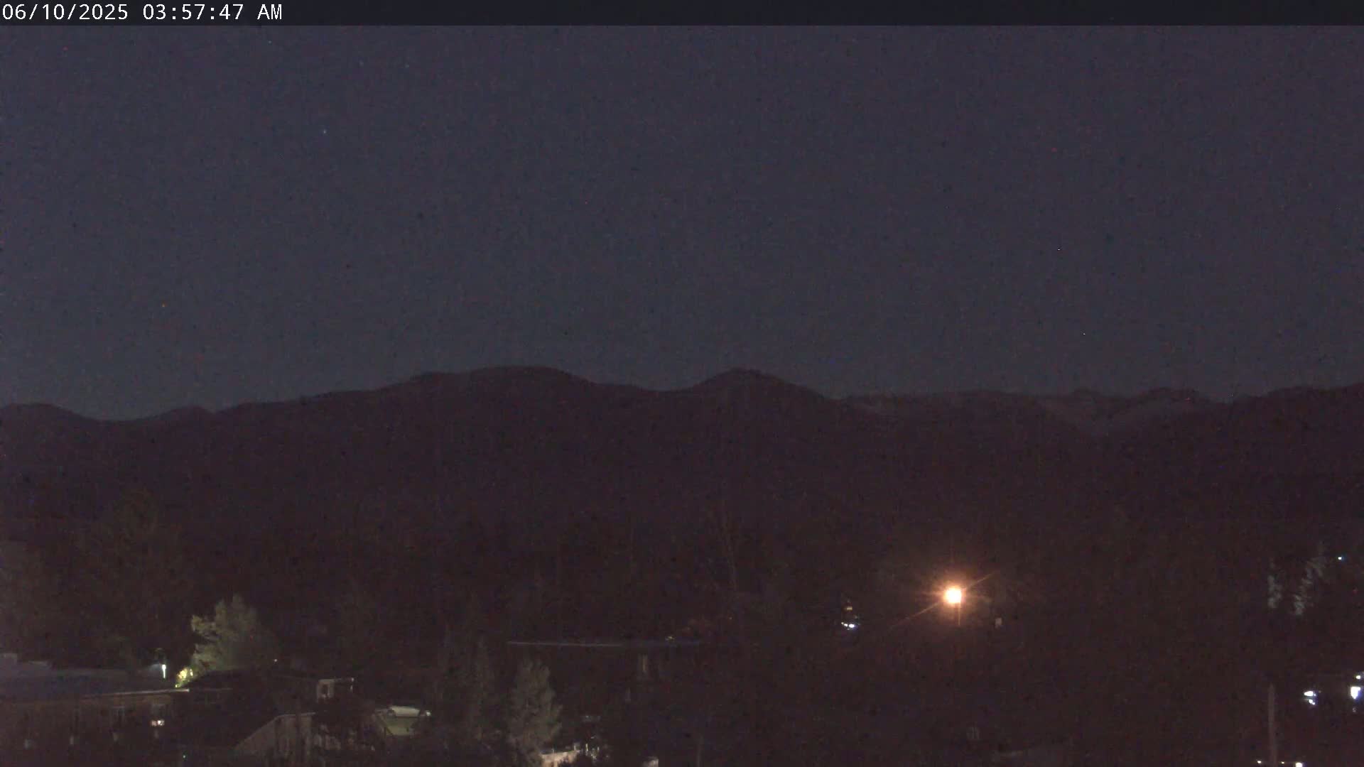 A dark, nighttime scene shows a mountain range in the distance, silhouetted against a clear, dark sky, with some lights visible in the foreground.